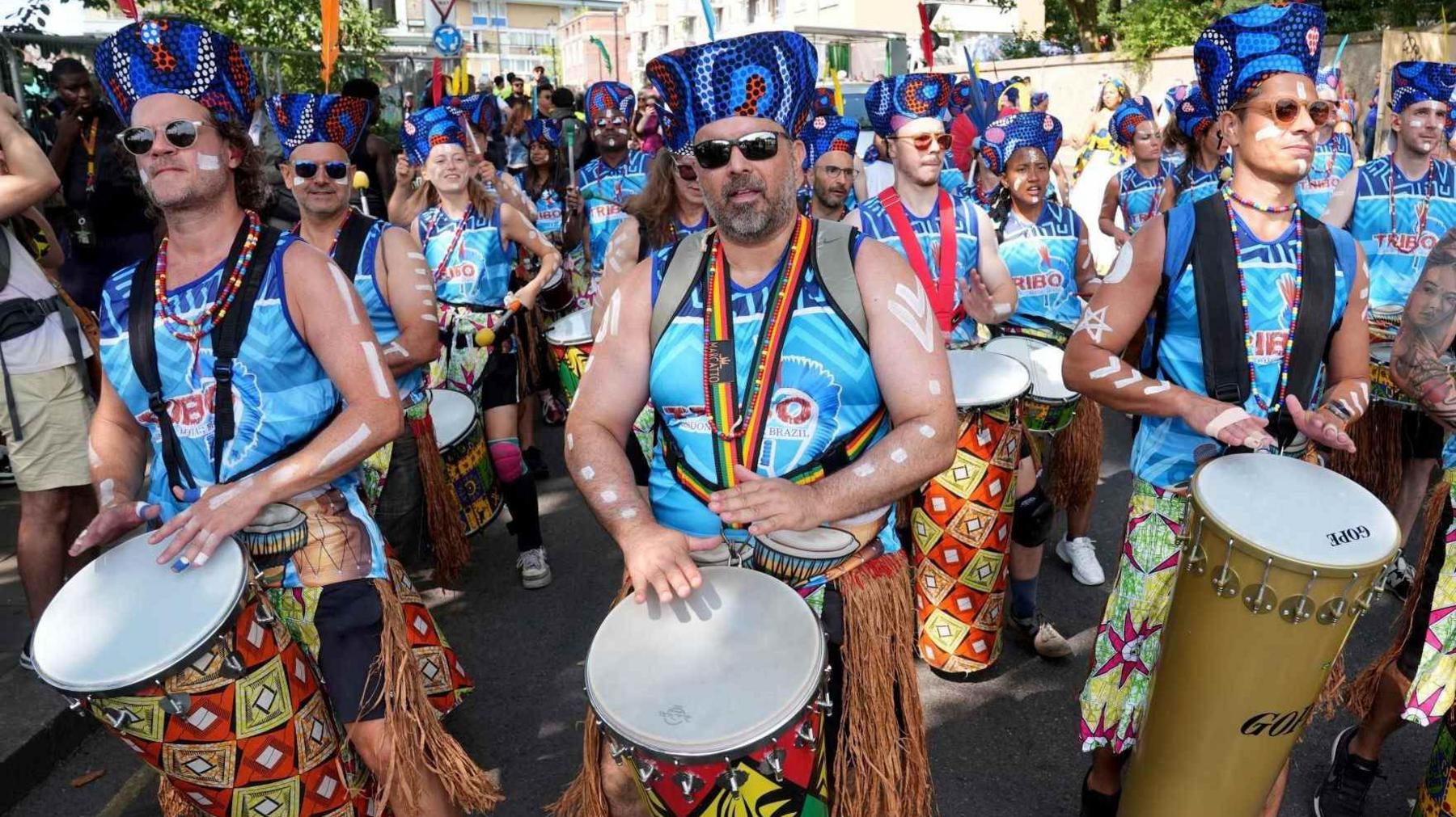 A marching band playing the drums with white body paint and blue hats