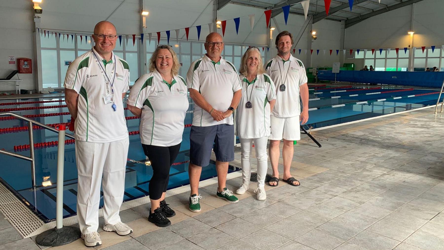 Five swimming coaches are standing next to each other by a pool, in the middle stands Mark Podbury. They are wearing a white coach unform and smiling at the camera. The pool is empty.