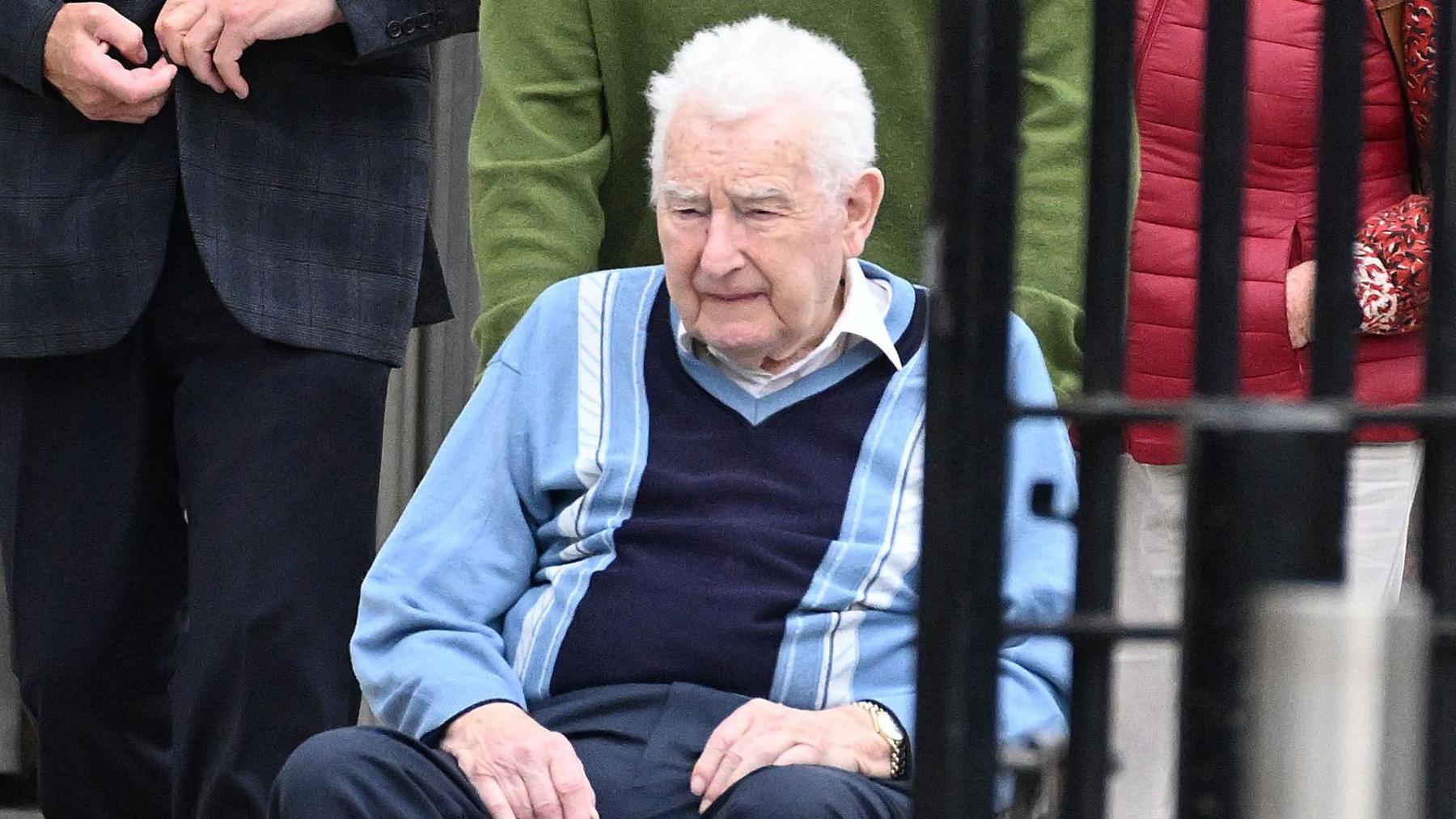 An elderly man with white hair in a wheelchair wearing a navy and blue striped jumper