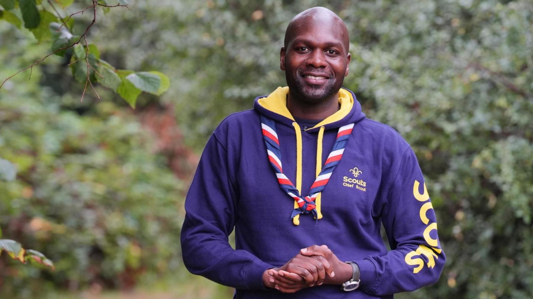 Dwayne Fields, who has been appointed chief scout. He is wearing a purple hoodie and is surrounded by trees. He is standing with his hands clasped together and smiling into the camera. 