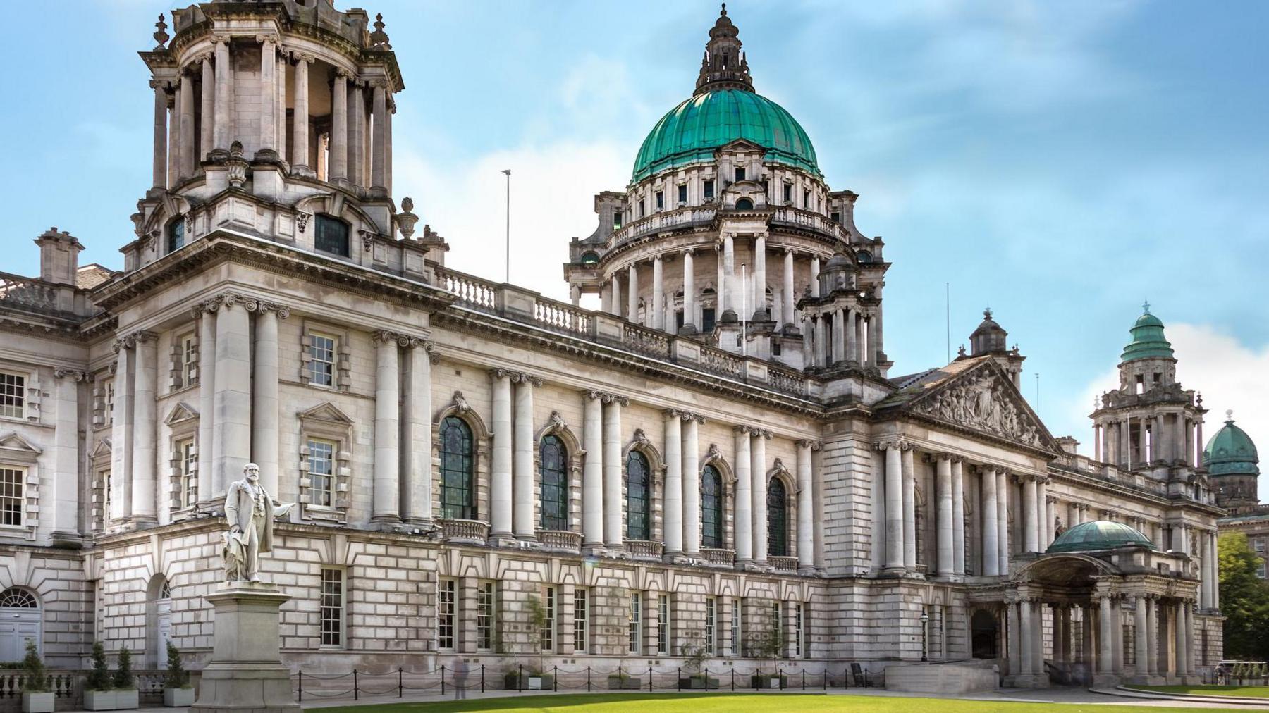 A grand looking white washed building with pillars, beautiful blue stained window glasses and deep panes and a teal blue roof with spires.
Outside on the grounds grass is visible and there is a statue of a man standing on the right.