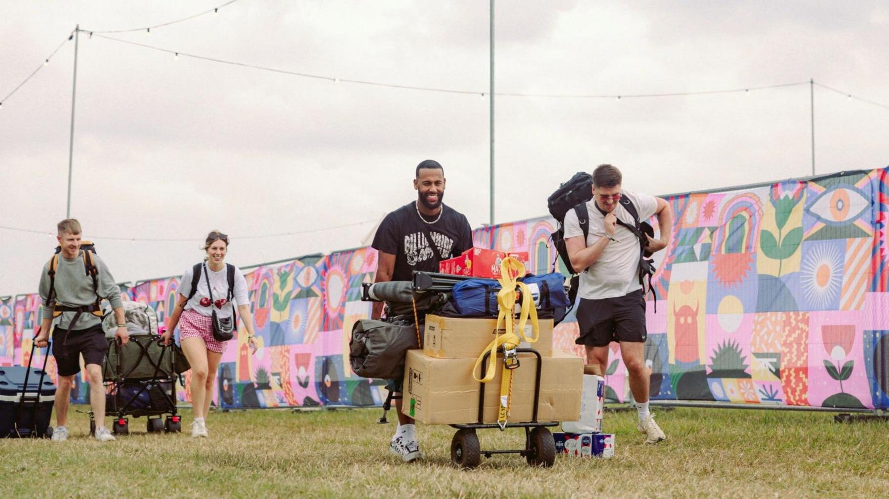 Four festival goers arrive at the venue, there are all wheeling trunks with their camping gear and luggage, smiling. There is a colourful backdrop sign lining the festival grounds in the background.