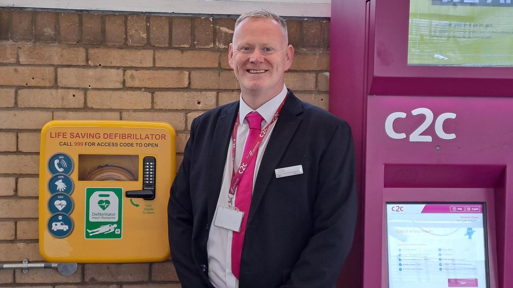 A man in a suit smiling in front of a yellow defribrillator box and a purple ticket machine