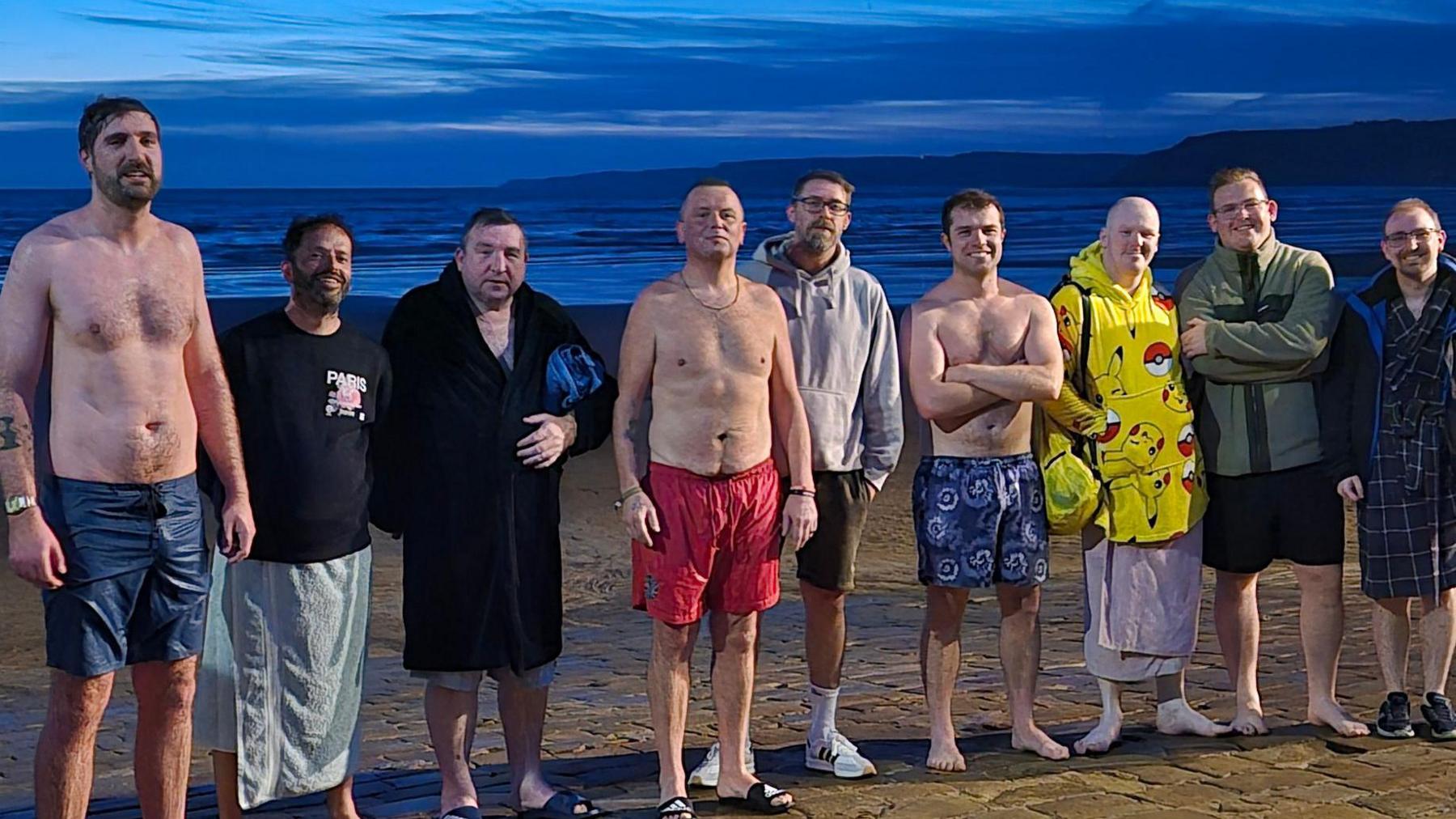 Group of nine men at South Bay beach in Scarborough on a summer's evening, with some in bare feet, some in trainers and sliders, and wearing towels or shorts