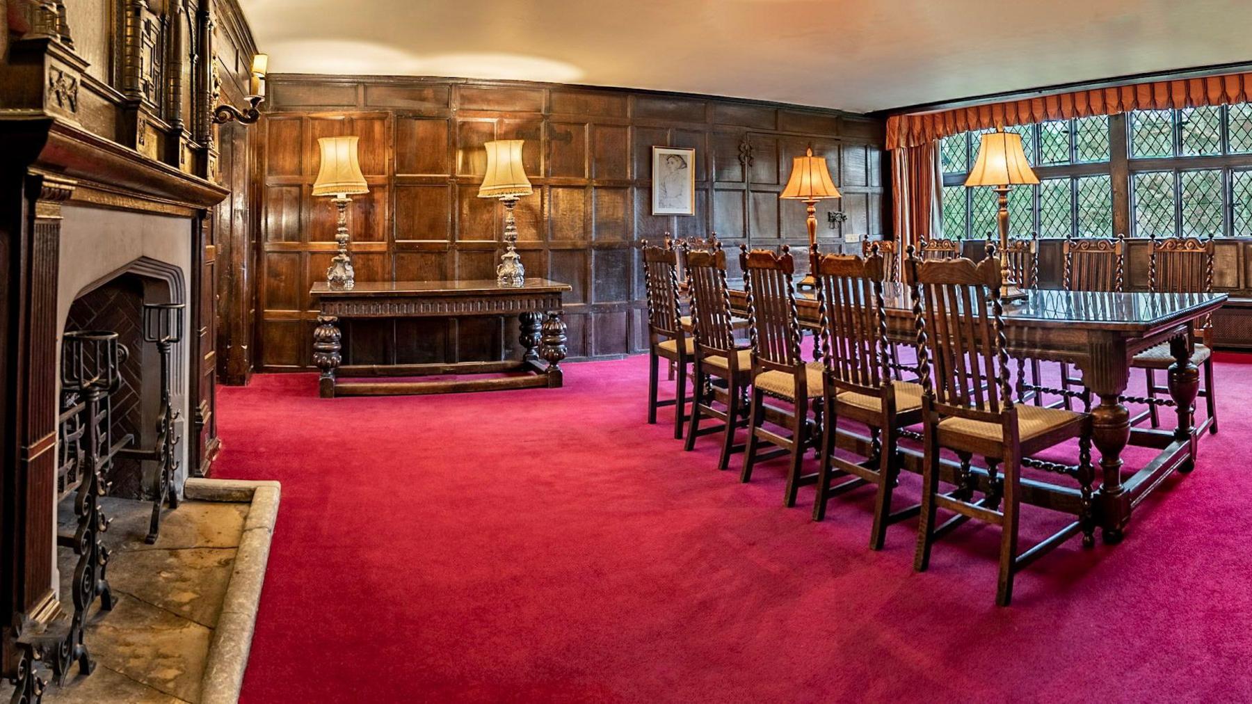A wood-panelled dining room with a bright red carpet. There is a long wooden dining table with five chairs on each side, four antique lamps, and a large fireplace furnishing the room.