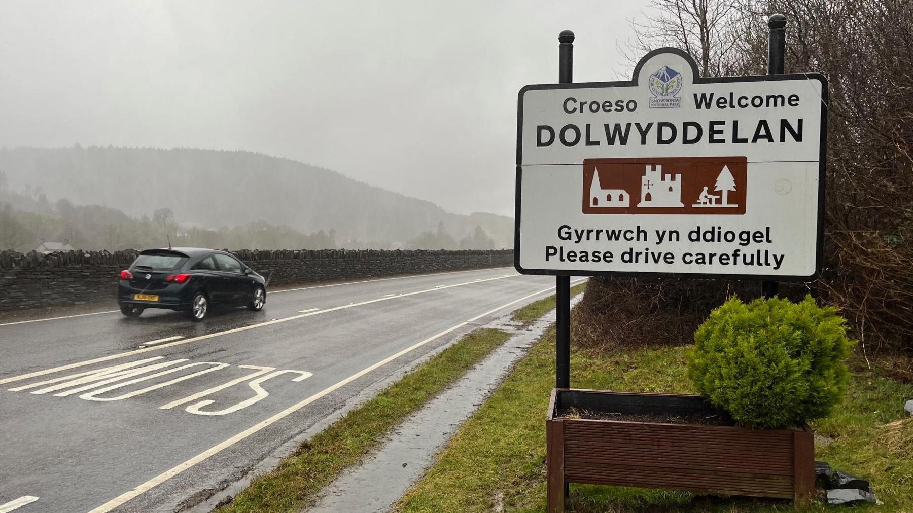 A bilingual sign at the edge of the village of Dolwyddelan with A470 to the left of it and a black car passing.  It's raining hard.