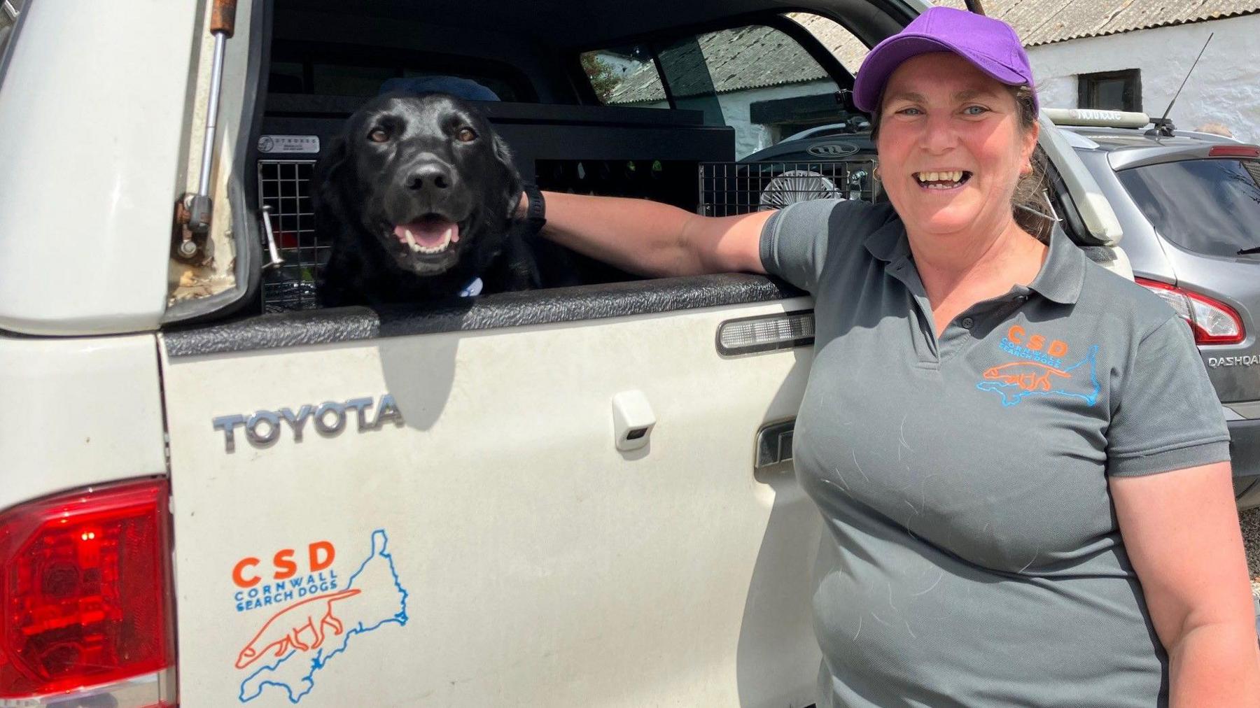 Annie Page is smiling at the camera as she stands at the back of a white pickup truck. Black labrador Lyla is sitting in the back of the truck and looking through the open window. Ms Page is wearing a baseball cap and a branded T-shirt and has her long brown hair tied back. On the back of the truck is the same as the logo on her T-shirt - it reads CSD CORNWALL SEARCH DOGS and has a line drawing on a search dog overlaid a line drawing of Cornwall.