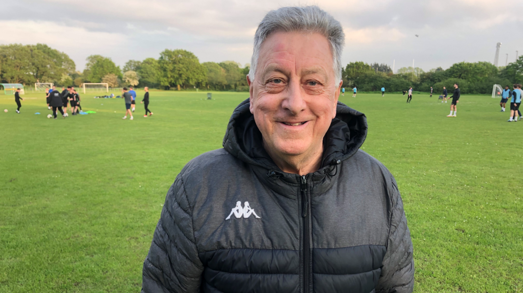 David Patient, who is wearing a black coat and standing in front of people playing football