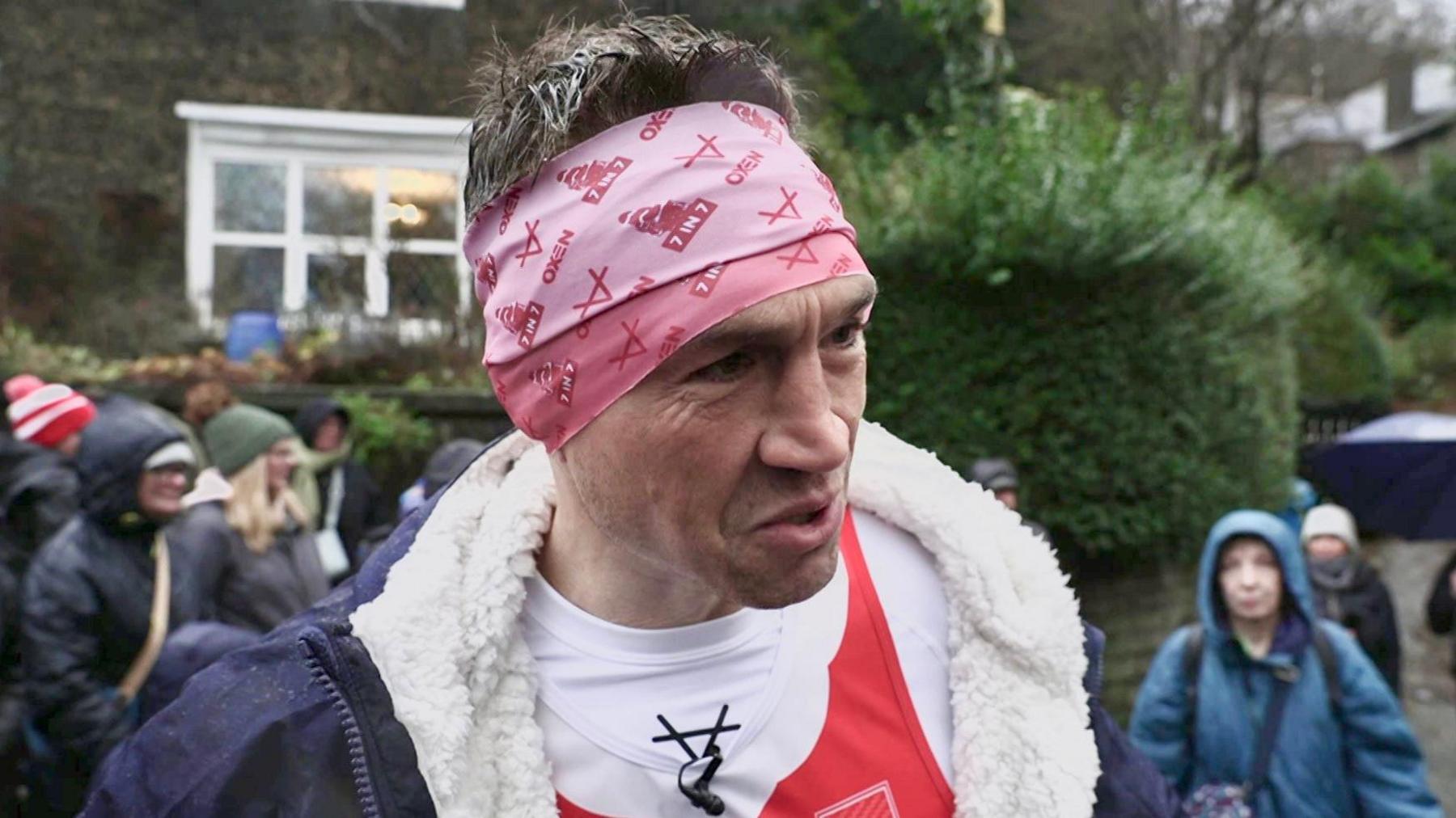 Sinfield, wearing a pink headwrap, speaking while surrounded by well-wishers lining a Saddleworth road