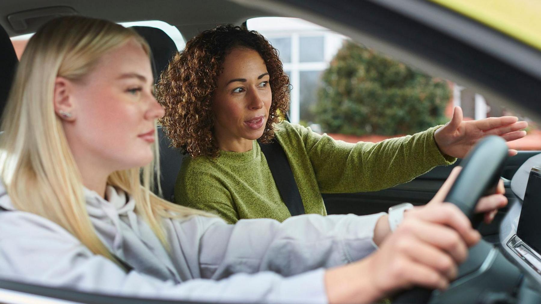 Young blonde woman sits in the drivers seat of a car as she listens to instructions from a female driving instructor