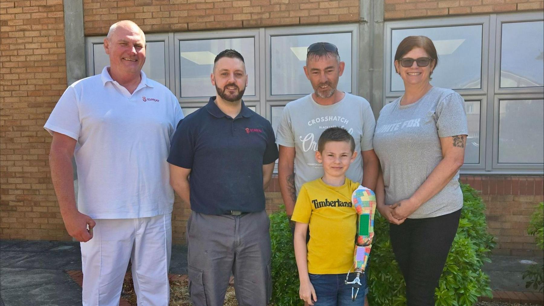 Three men, a woman and a young boy with a prosthetic arm standing in front of a building and smiling at the camera.
