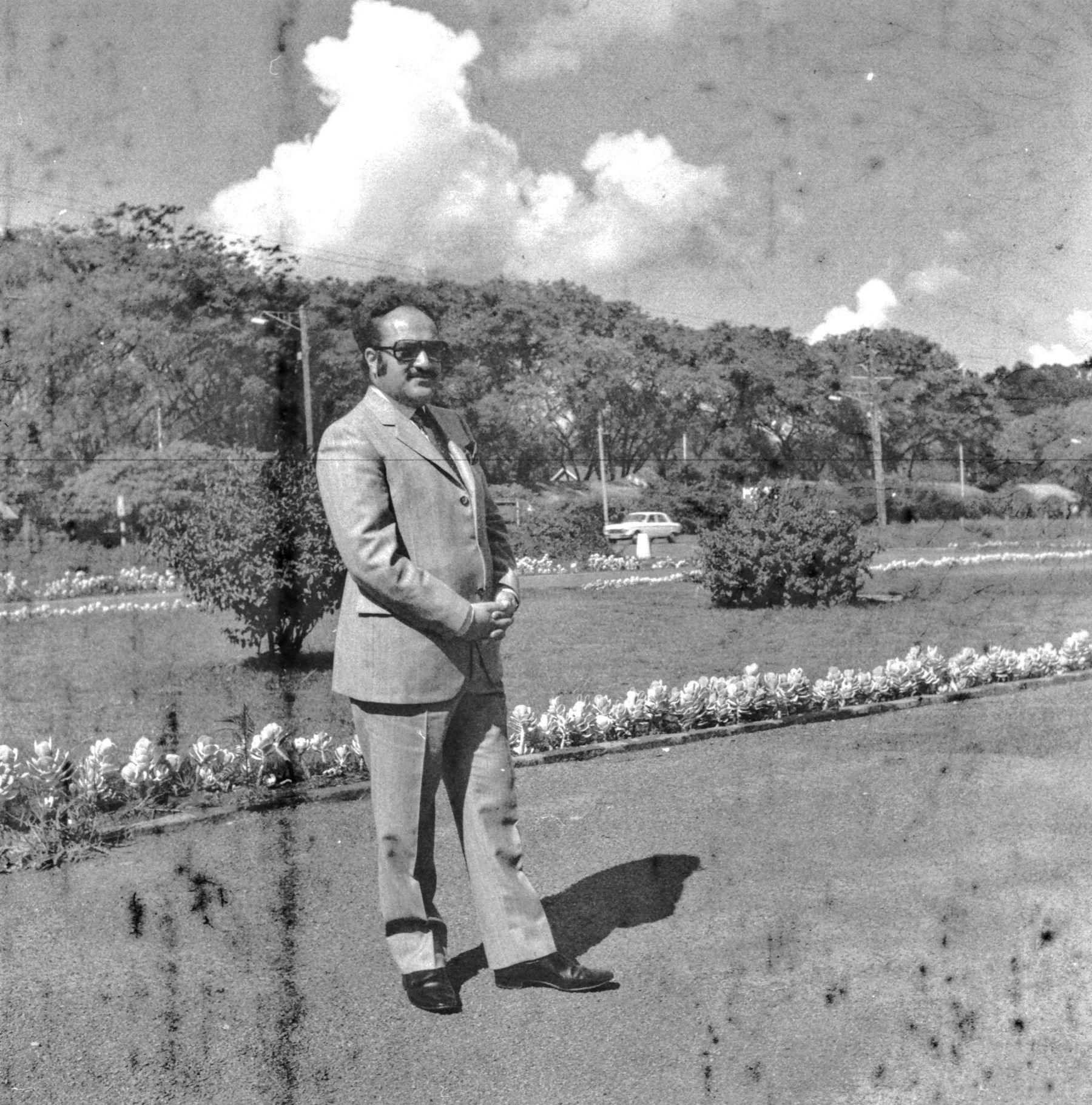 A black-and-white photograph of Susheel in a smart suit. He has a moustache and is wearing sunglasses. There is a garden with flowers and trees behind him.