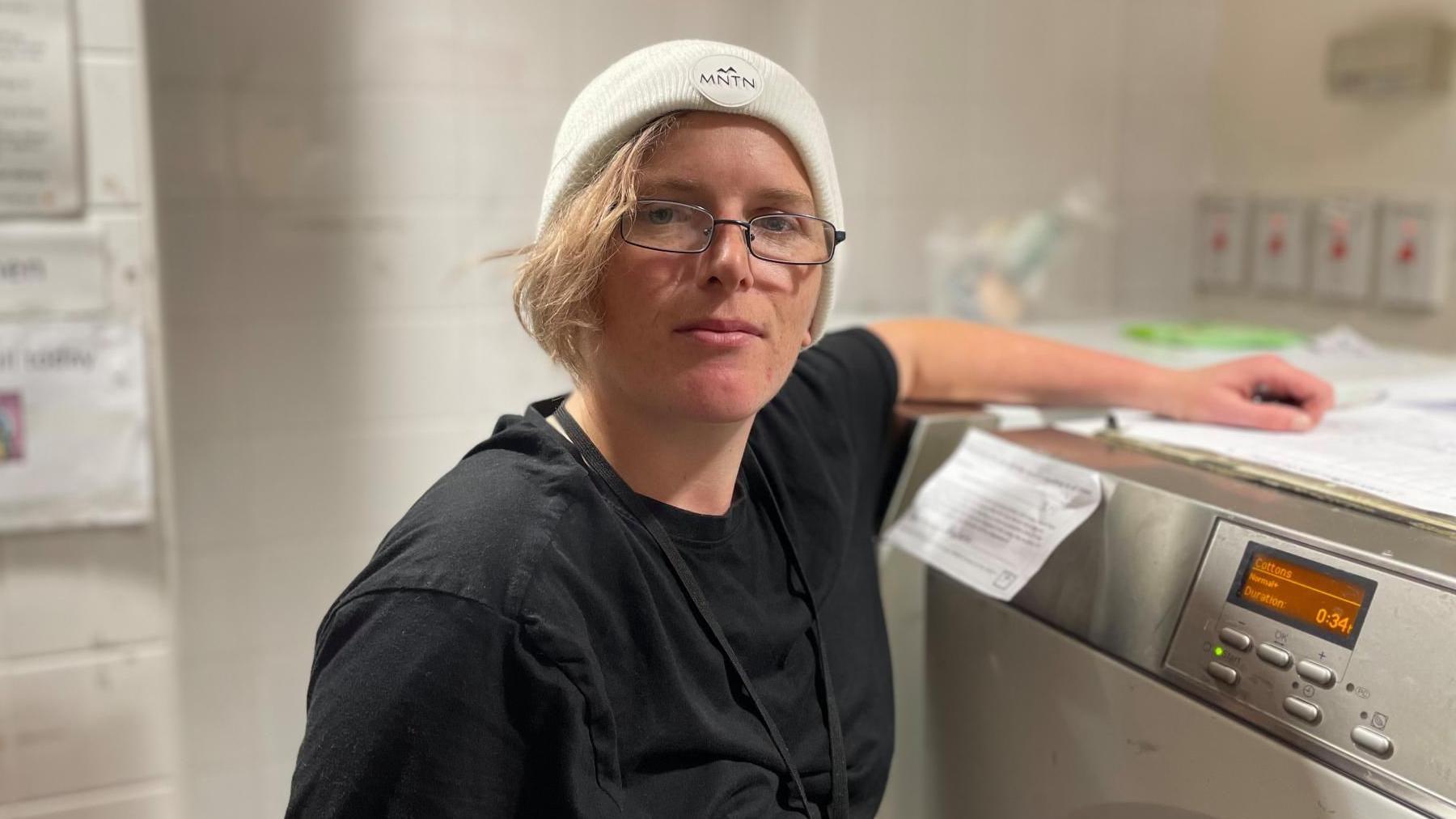 A young woman in a black t shirt and grey hoodie stands next to a washing machine