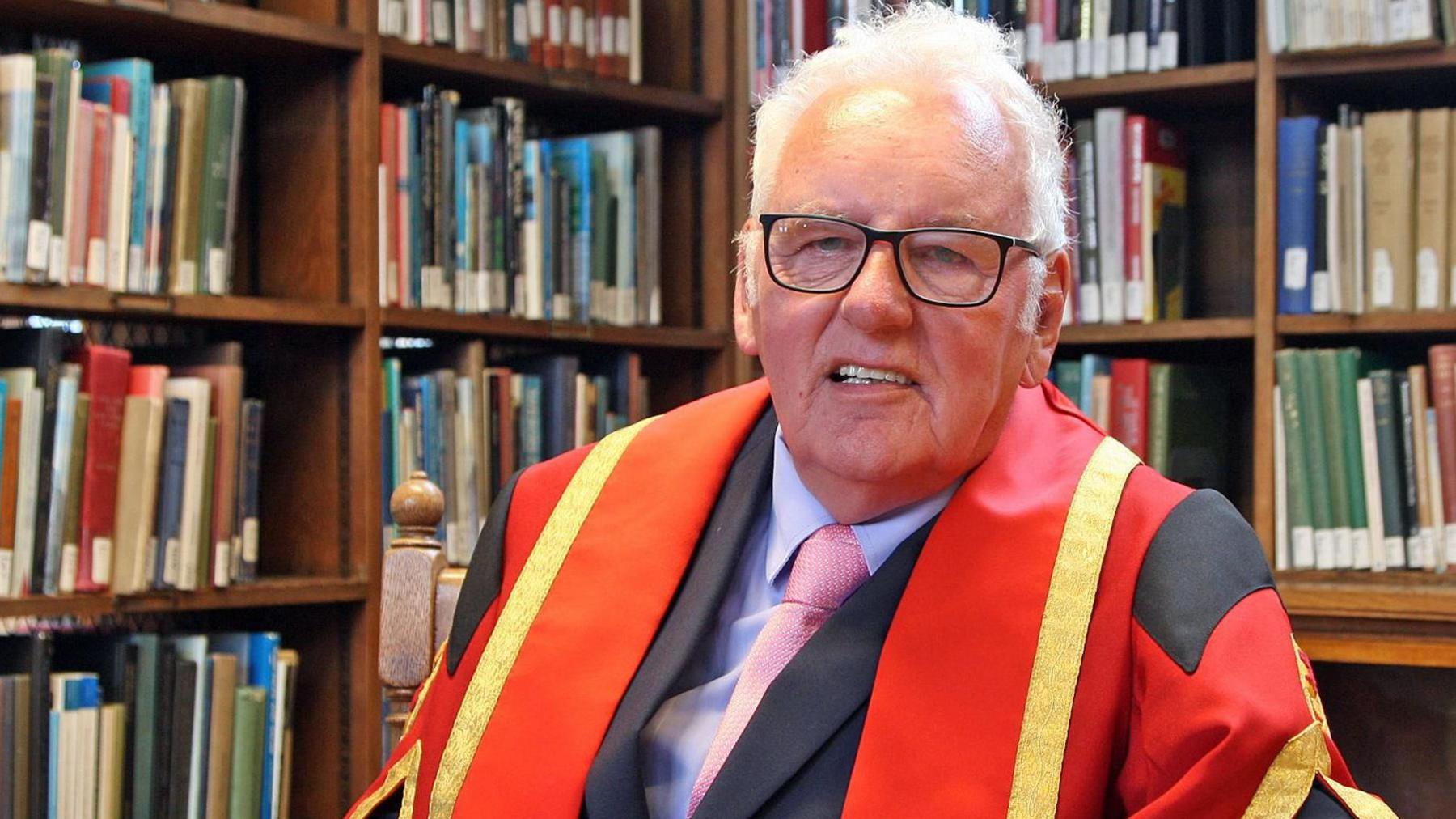 Noel Thomas in his graduation gown, sitting at a wooden table in front of bookshelves