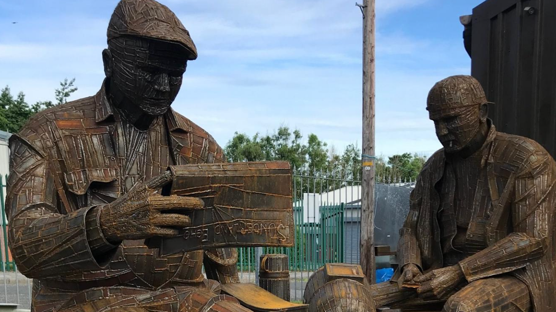 Two sculptures of men - one is wearing a flat cap and reading a newspaper. The other is wearing a helmet.