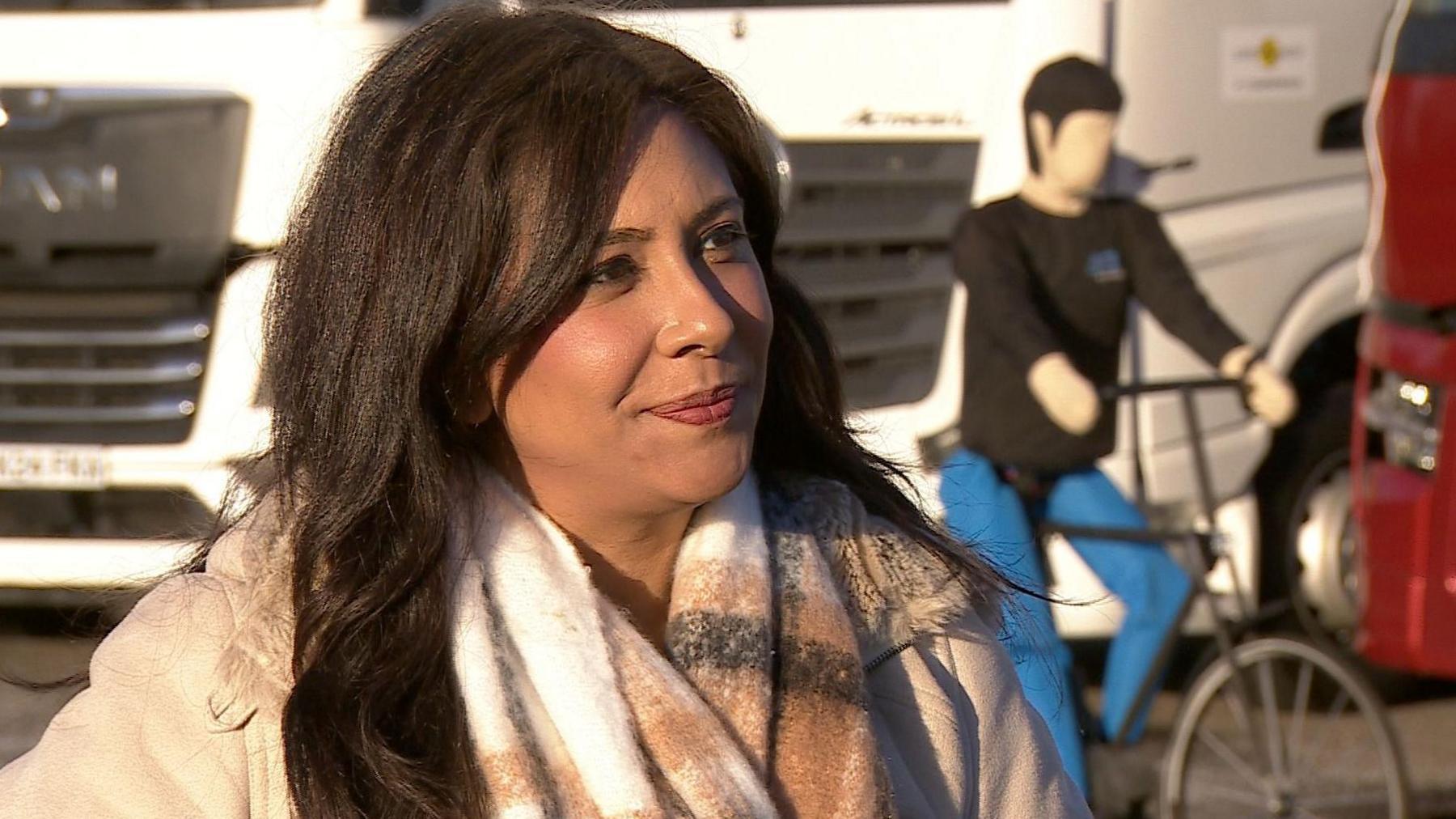 Meera Naran in a light brown coat and scarf stands in front of a lorry and test dummy on a bike.