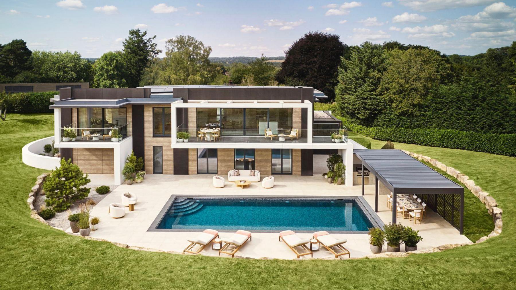 A large luxury house with a swimming pool in front. The property is surrounded by grass and trees in the background