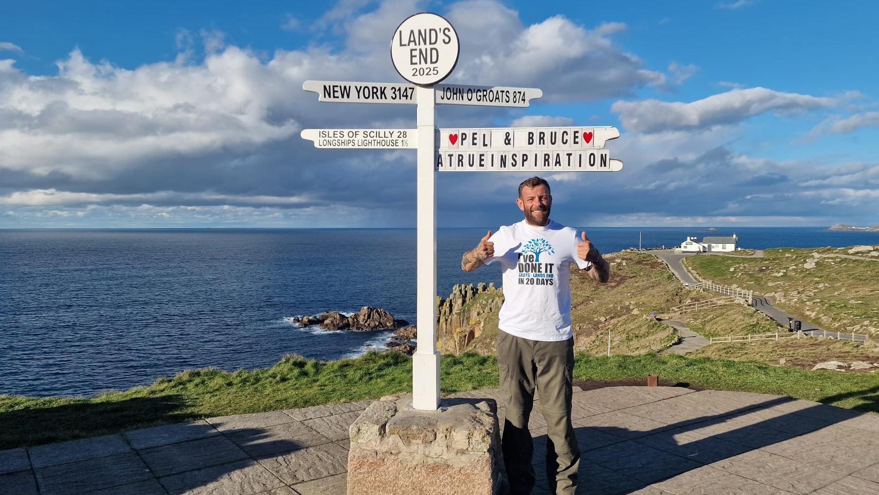 Perry Pel Scopes stands in front of the Land's End sign. The sea is in the background and the sky is blue. He is wearing a white tshirt and brown trousers. The sign says 'Pel & Bruce a true inspiration'