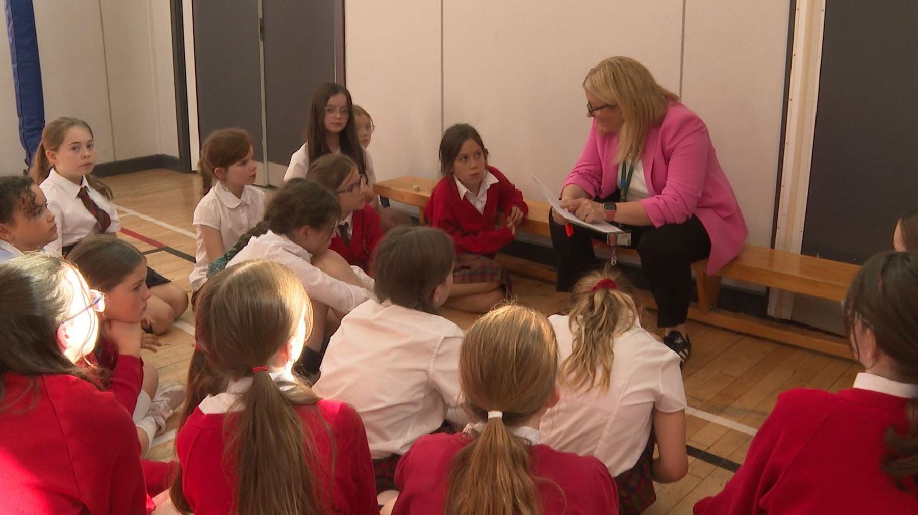 Boireannach bàn le speuclairean oirre le seacaid phinc, briogais dhubh na suidhe a' bruidhinn ri buidheann de chloinn-sgoile air an èideadh ann an deiseachan-sgoile - lèintean geala agus geansaidhean dearga