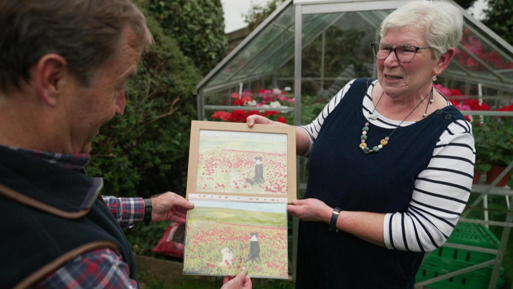 David Braine and Julie Taylor look at a framed painting featuring a black and white dog and a blond dog looking out at a filed of red poppies, and her photograph of the same scene. 