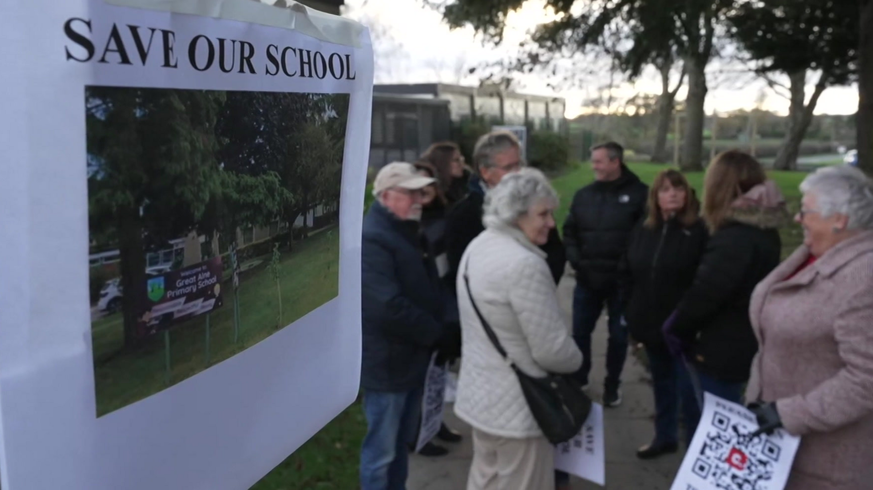 A poster saying "Save Our School" is on the left of the image with a group of men and women standing outside the school on the right. They are near some tall trees which can be seen behind them and school buildings are also on the left.
