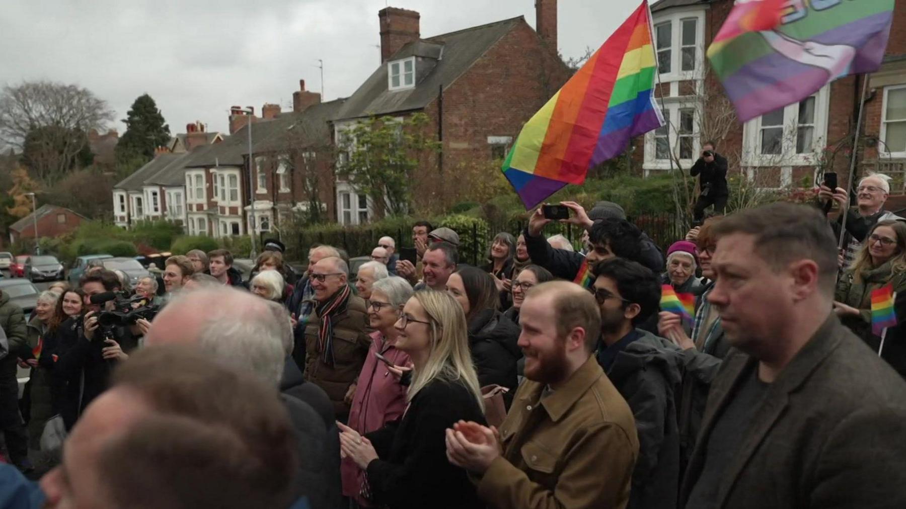 A large crowd of smiling men and women gather to watch the unveiling. They are standing in the road and there are houses visible behind them. Many are clapping, some are wearing colourful scarves and waving multi-coloured flags.