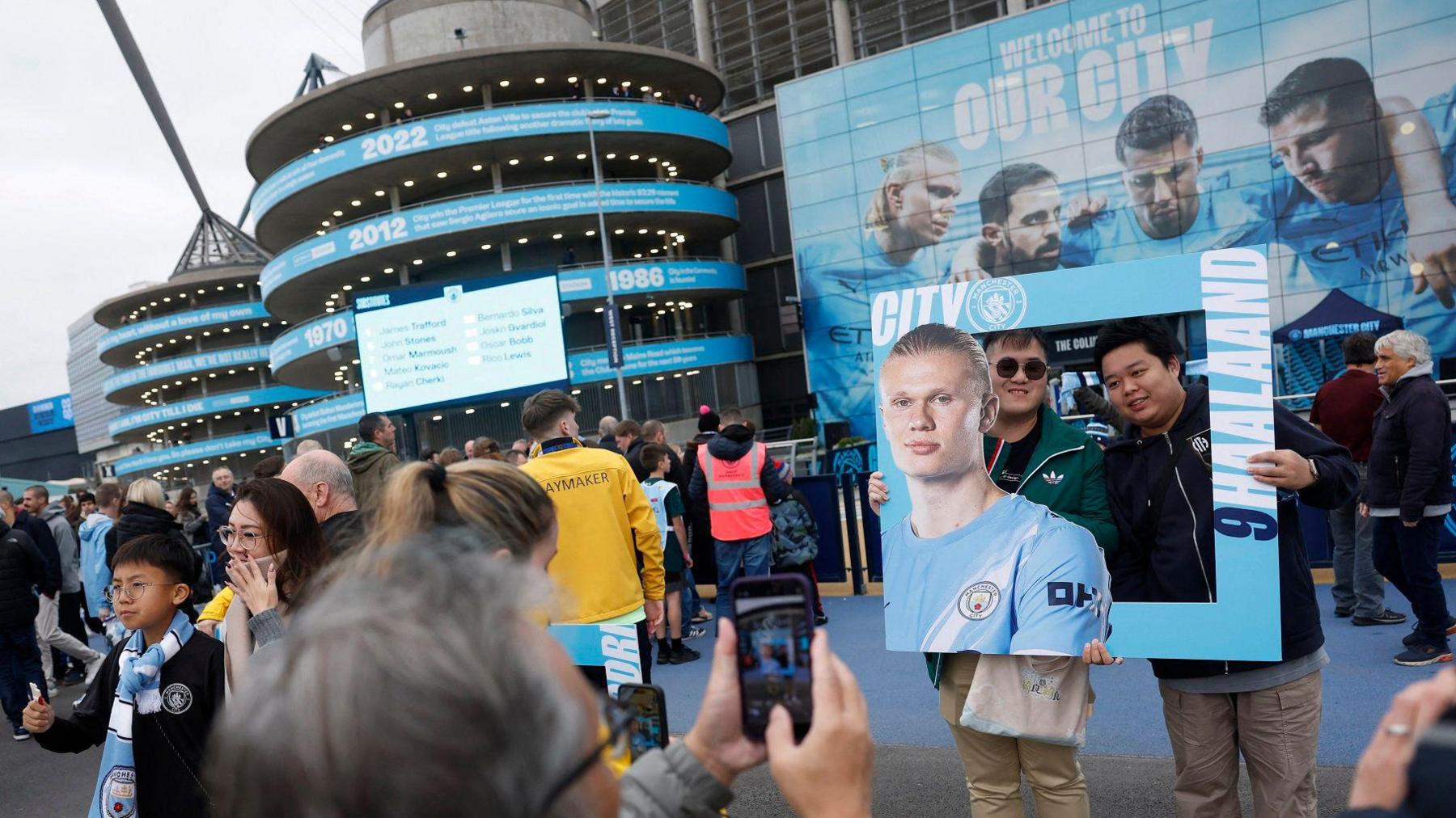 A man takes a picture of two fans posing in a frame they're holding, which shows a picture of Erling Haaland. They are outside the City stadium on a matchday with fans milling around in the background.