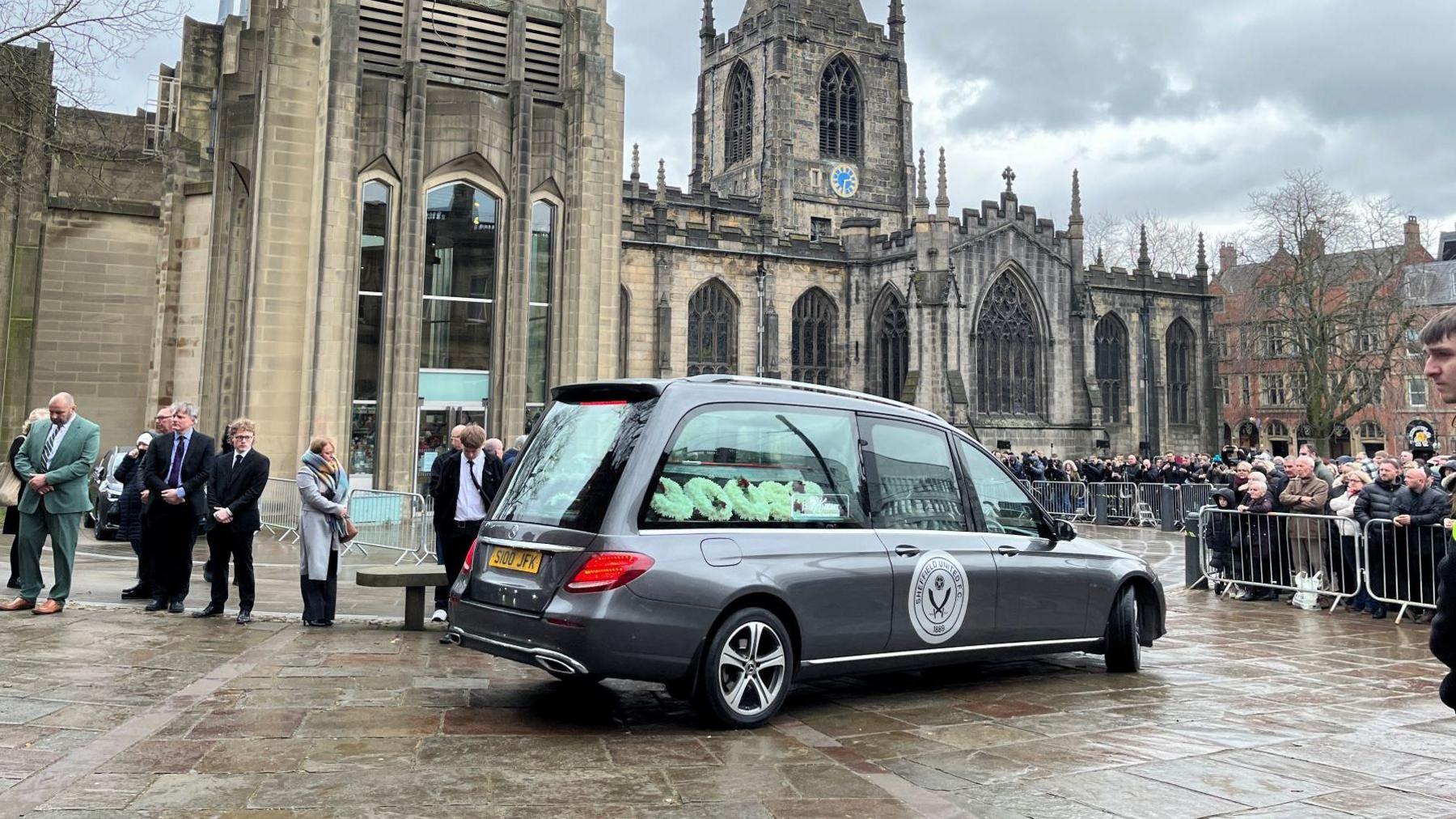 A grey hearse with a Sheffield United logo parked outside Sheffield Cathedral. A floral tribute saying Goose can be seen inside the vehicle as crowds gather nearby 