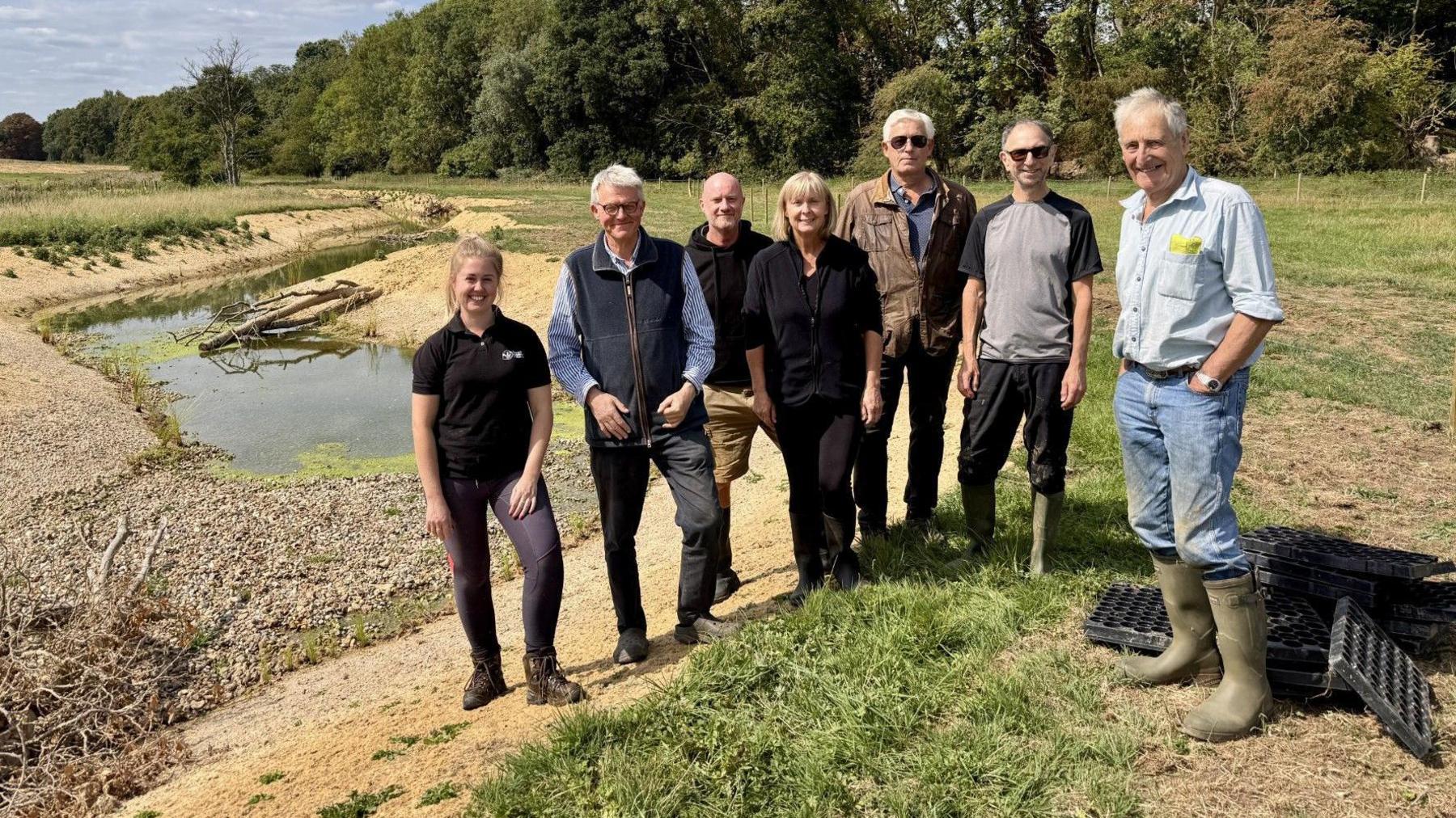 Seven people, looking at the camera, and standing on a bank, by a river. There are two women and five men. They are all wearing trousers. 