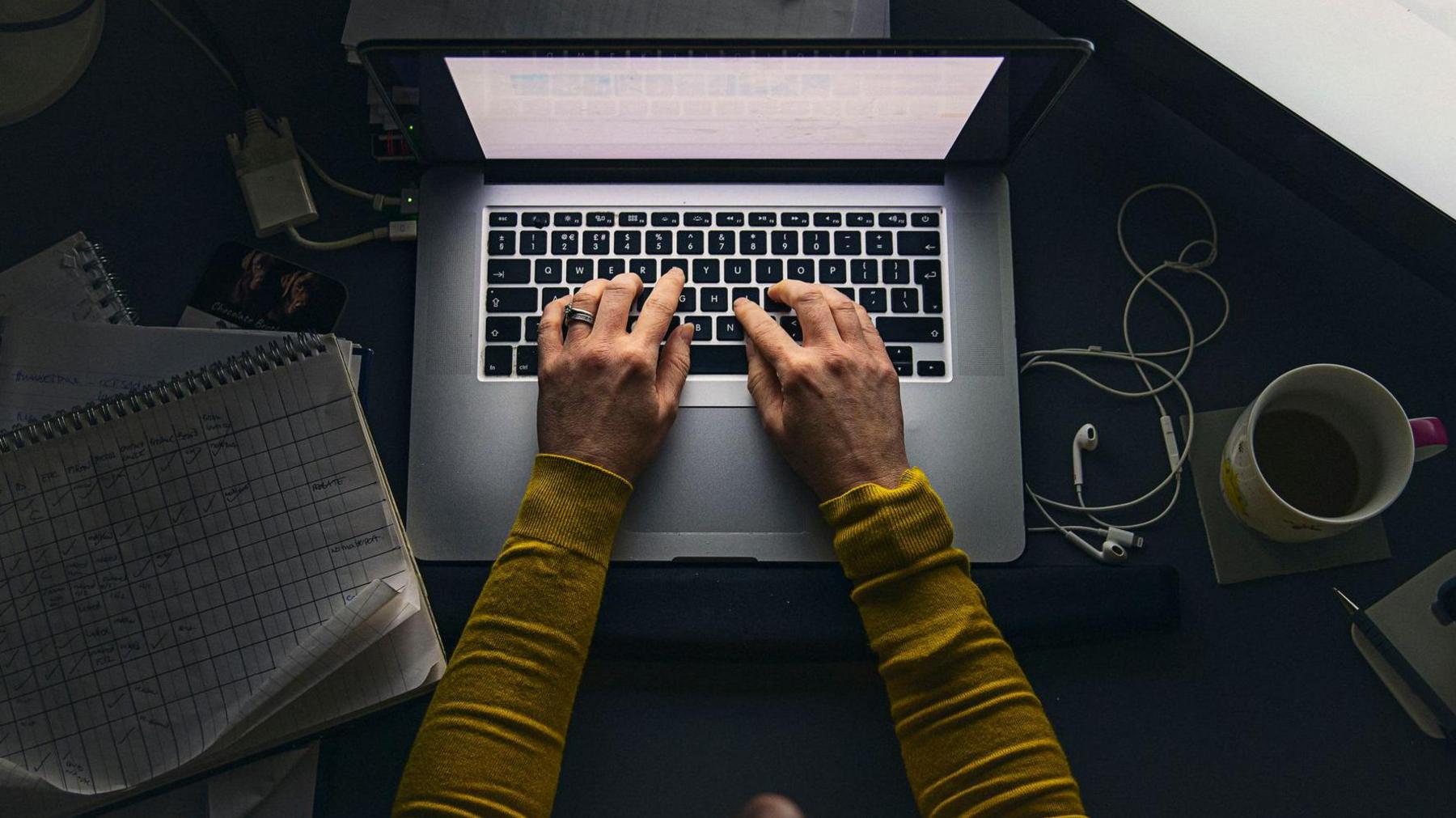 A woman is sitting at a desk and is typing on the keyboard of a laptop. There is a set of small headphones and a coffee cup next to the computer. A pad of paper with a grid drawn on is also on the desk.