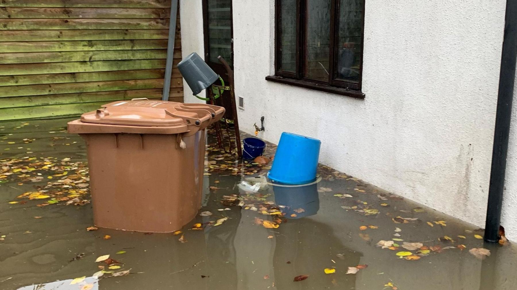 Dirty water half way up the wall of a house and a brown wheelie bin. There is a bucket and lots of brown leaves floating in the water.