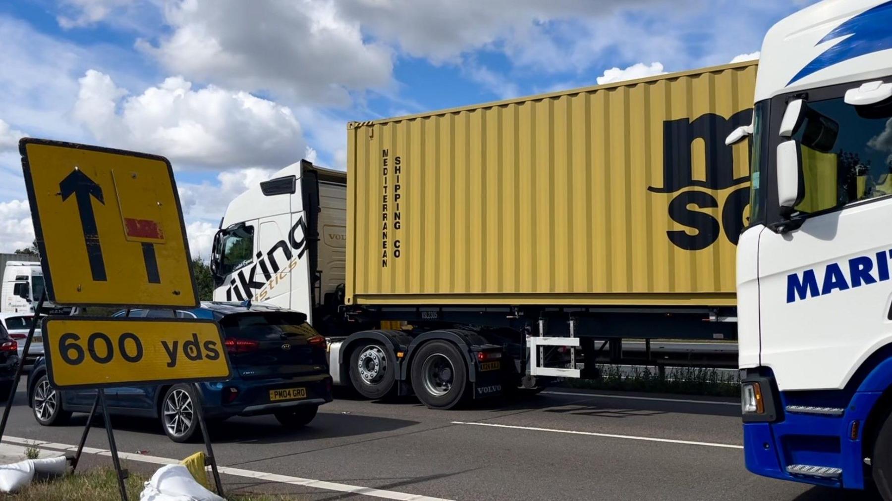 Traffic on bridge, including a yellow lorry and a blue kia, with a lane closure warning sign in the foreground