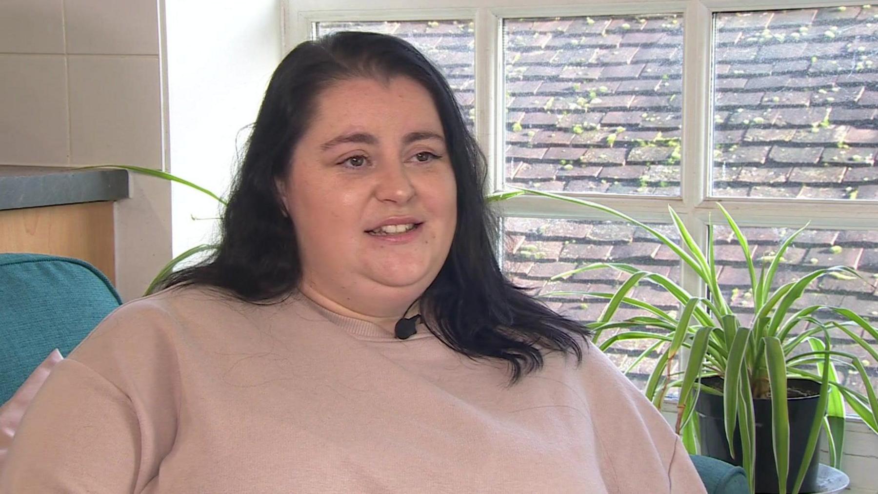 Rosie sitting in front of a window. She has black hair and a light brown jumper, and is looking slightly away from the camera. 