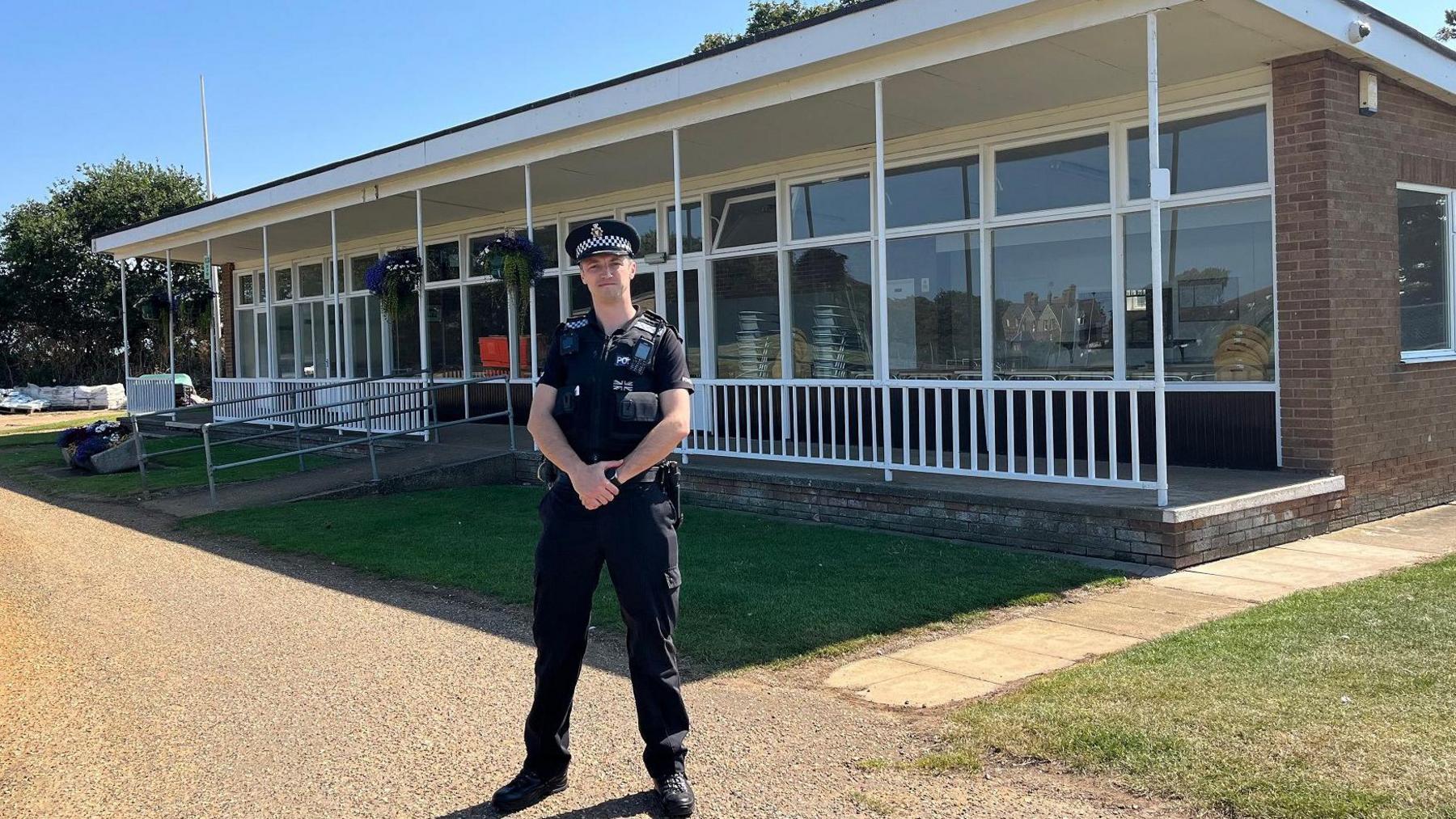 Police inspector Ben Jarvis is wearing a black uniform with a short sleeved shirt because it's a very hot day. He is wearing a police hat. He is standing in front of the club house at Hunstanton Recreation ground which is a one story building with pretty hanging baskets by the door. 