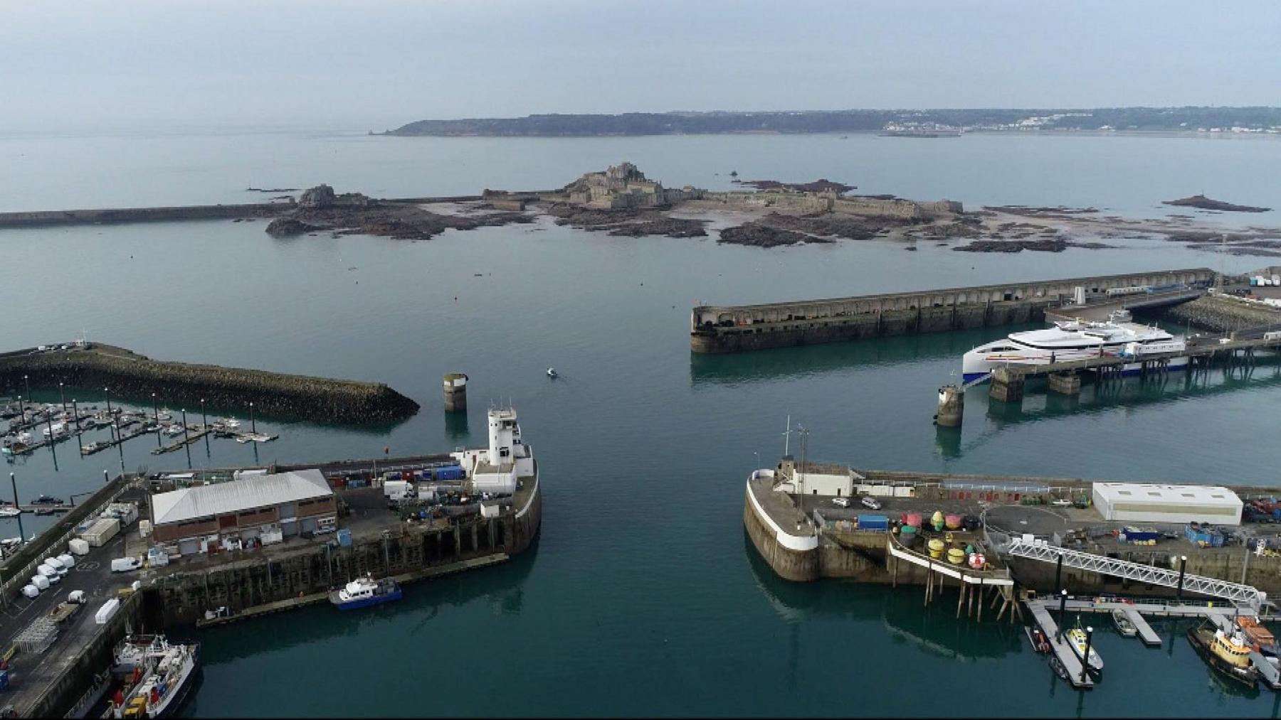 An aerial view of the Ports of Jersey - four separate walls sticking out in the water with boats surrounding, in the background you can see Elizabeth Castle and further afield is the coastline of Jersey. 