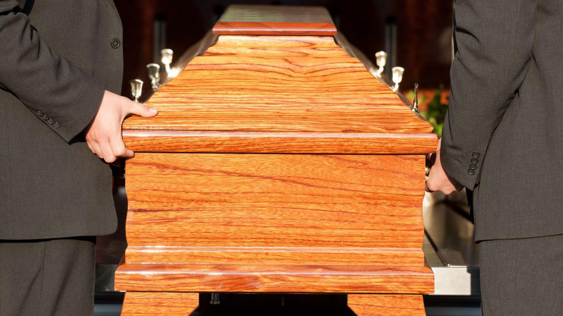 Two men wearing black suits stand either side of a wooden coffin. You can only see their arms and torsos.