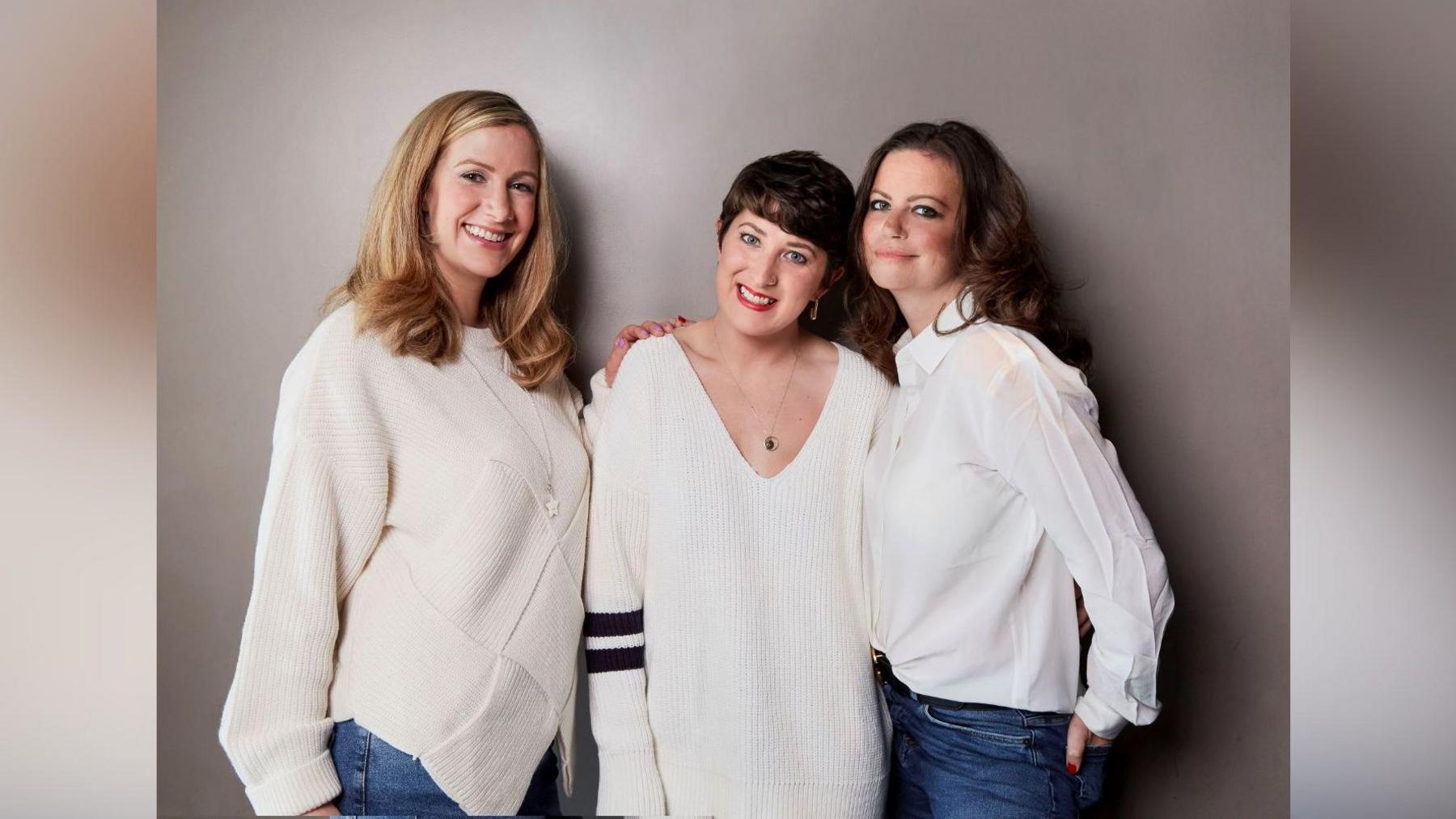 Formacioni origjinal i podkastit "You, Me and the Big C" të BBC-së. Nga e majta në të djathtë: Rachael Bland, Lauren Mahon dhe Dame Deborah James. Të treja po buzëqeshin para kamerës. Rachael ka veshur një bluzë të bardhë dhe xhinse, Lauren ka veshur një bluzë të bardhë me dy vija të zeza në mëngë dhe Deborah ka veshur një këmishë të bardhë dhe xhinse.