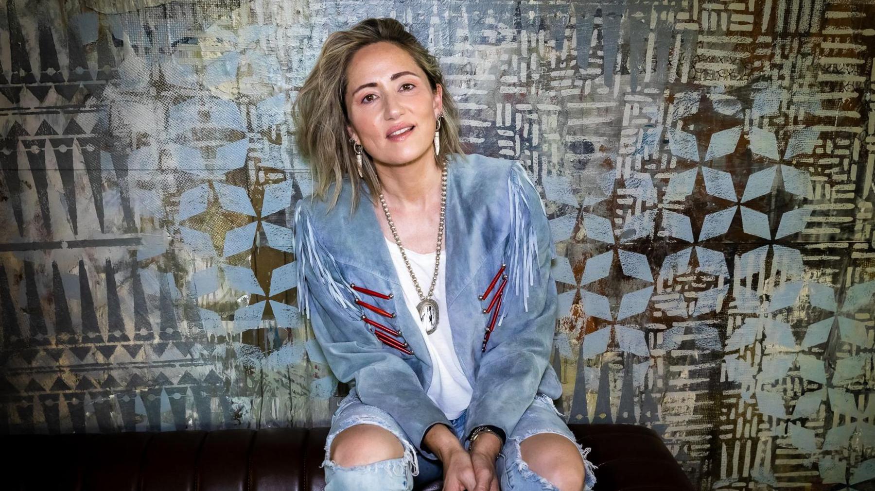 KT Tunstall in ripped denim jeans and a light blue suede jacket sitting smiling.