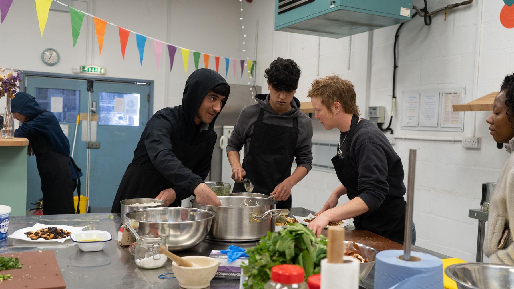 Three young people in black aprons are preparing food on a kitchen island. Pots and pans are on the island with ingredients.