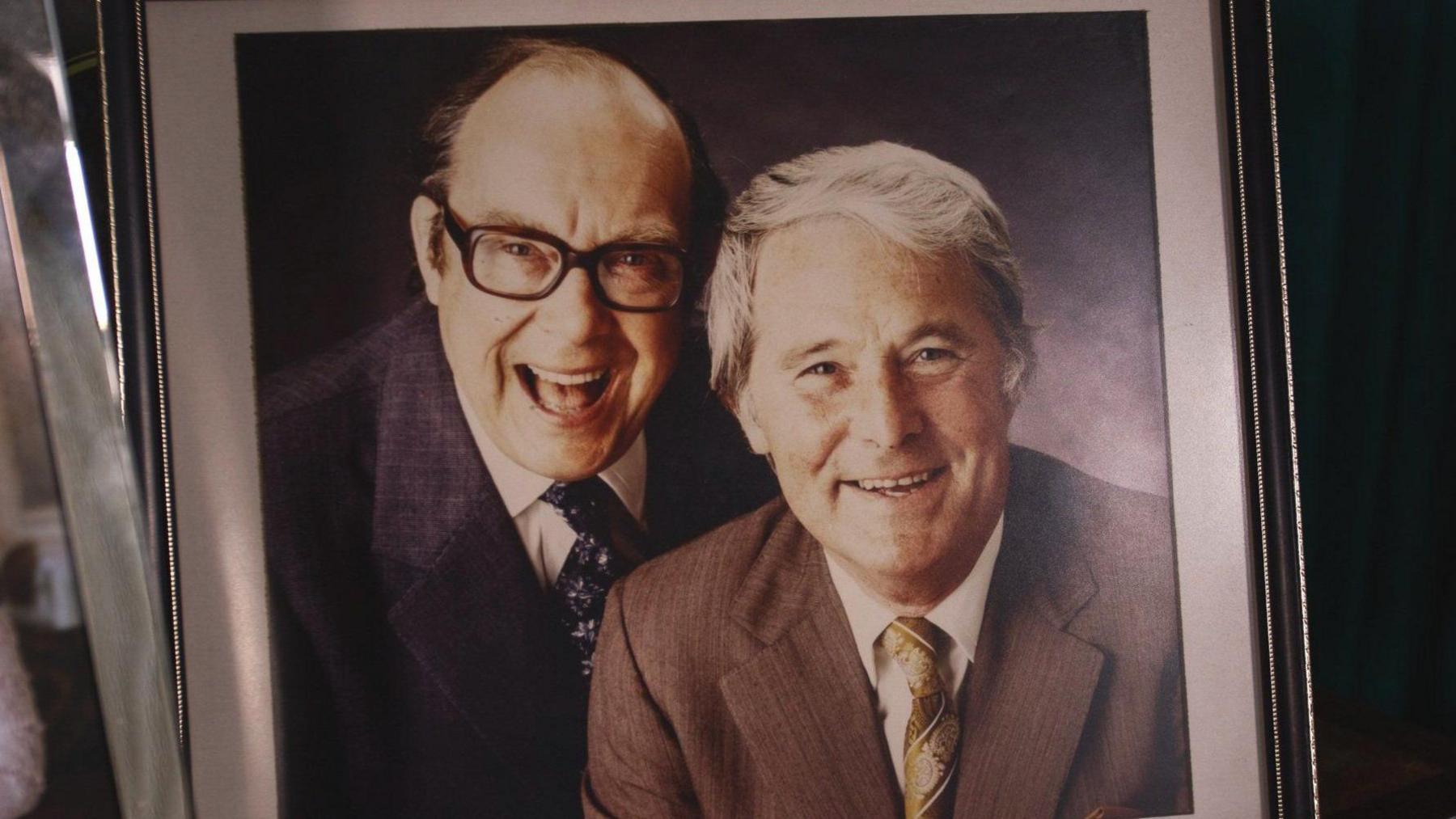 Eric Morecambe laughing at the camera with his signature, black framed glasses, with Ernie Wise to the right of him, smiling at the camera 