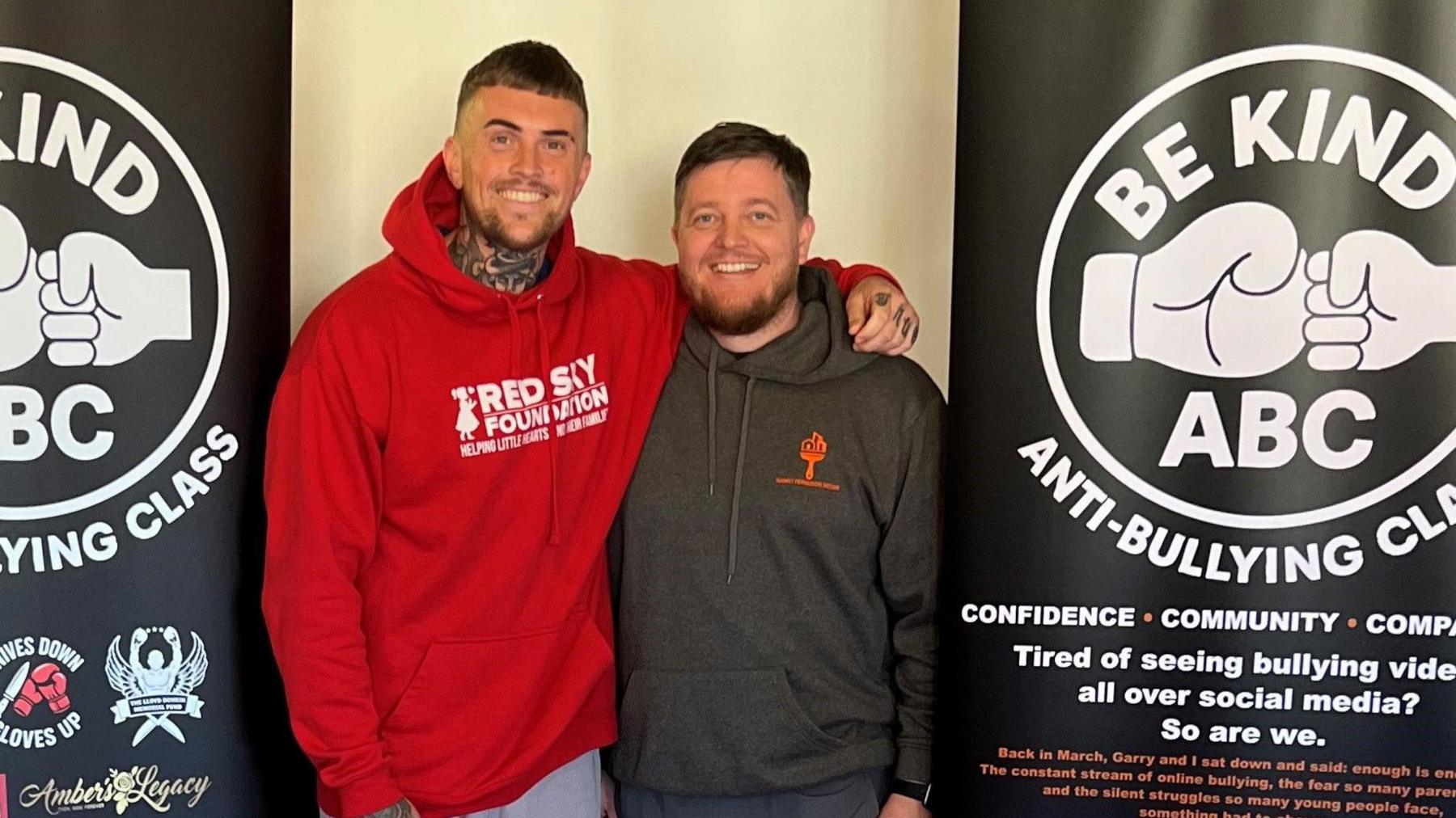 Troy Baker, left, has his arm around Gerry Ferguson's shoulder as they smile at the camera. Mr Baker is wearing a red hoodie with Red Sky Foundation written across it in white. Mr Ferguson is wearing a grey hoodie with an orange logo. They are standing between two black banners reading Be Kind ABC ANTI-BULLYING CLASS. the logo is of two fists, a larger one and a smaller one, presumably of a child, bumping into each other.