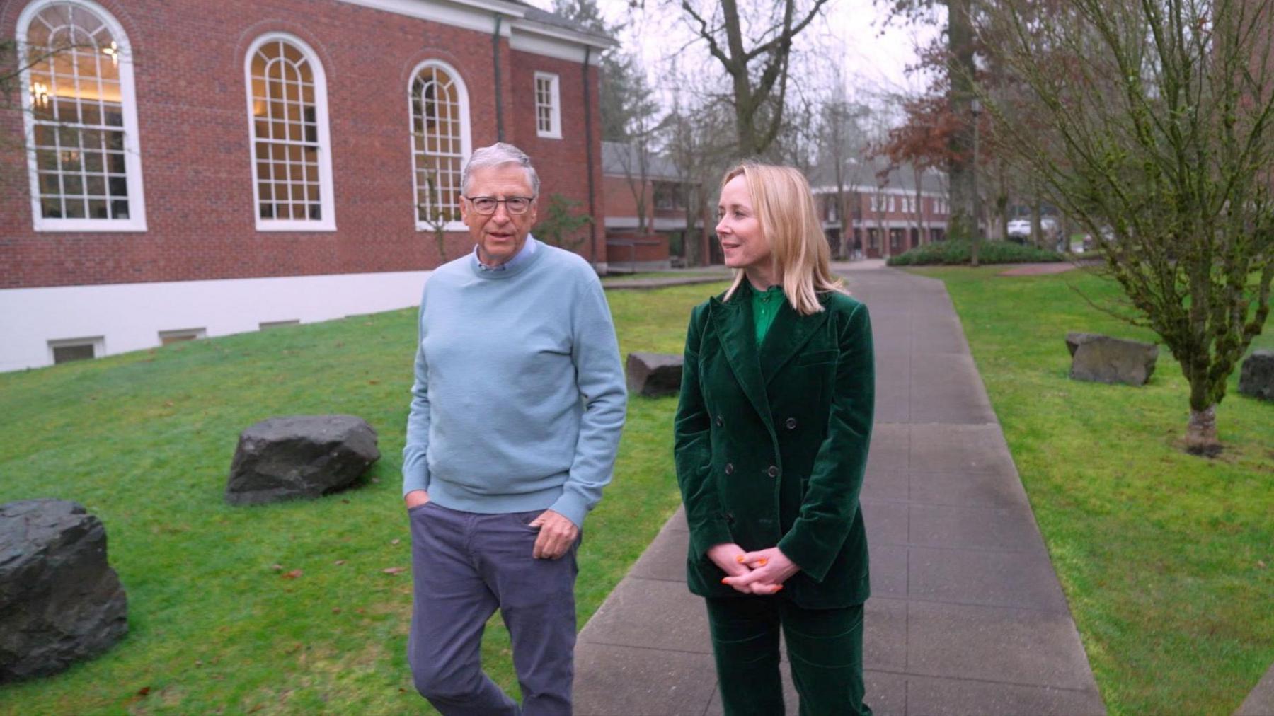 Bill Gates in pale blue jumper and grey trousers and Katie Razzall in velvet green trouser suit walking outside Lakeside School in Seattle 