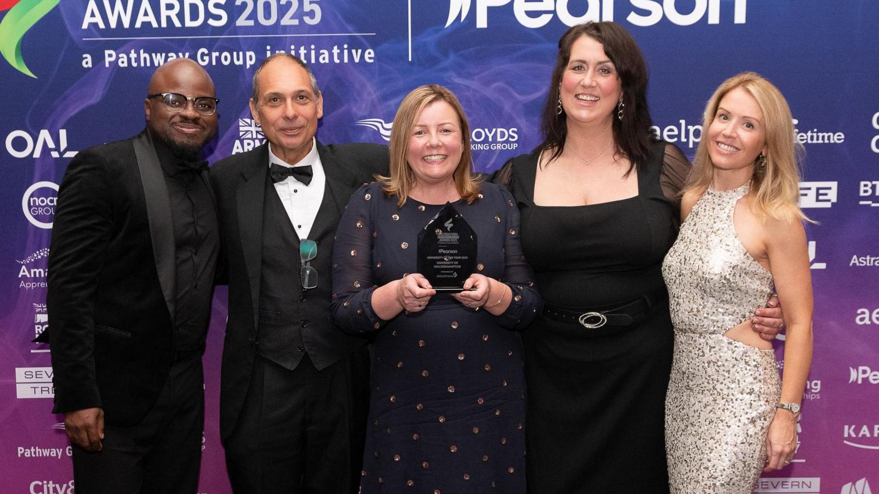 Two men and three women, dressed in smart outfits, smile as they stand in a line in front of a banner the names of sponsors over it. The woman in the centre is holding a small award with both hands.