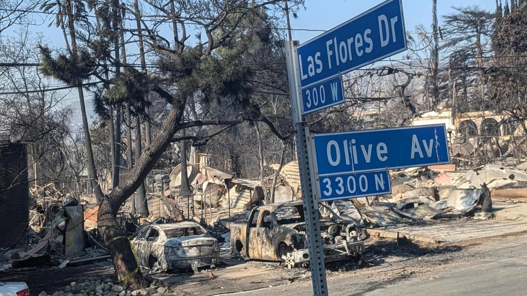 Street sign says Las Flores Dr and Olive Ave, with burned-out cars in background