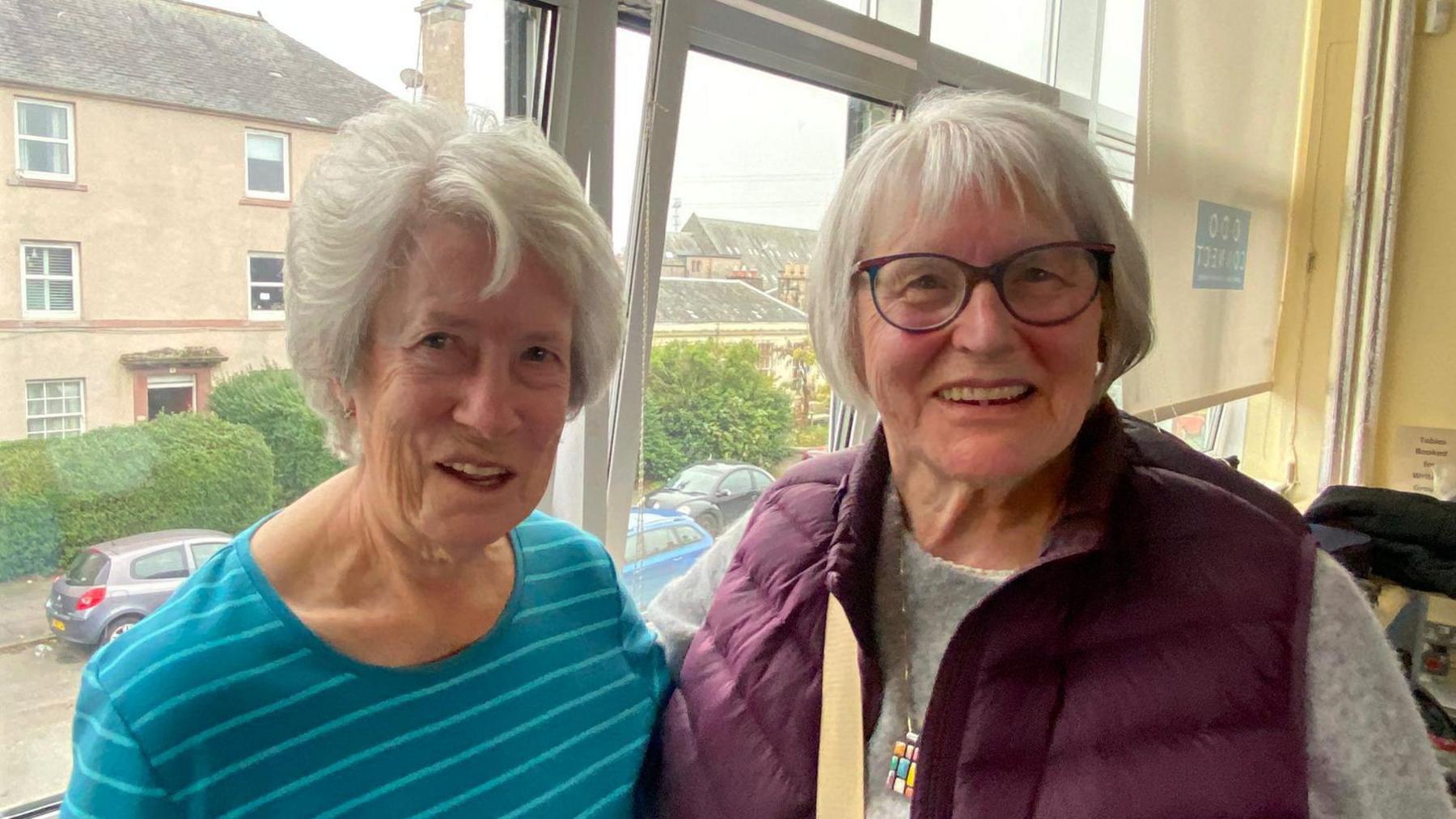 Two elderly women smiling at the camera. The woman on the left has short grey and is wearing a blue striped top. The woman on the right has short grey hair and glasses. She is wearing a grey jumper and dark purple gilet. 