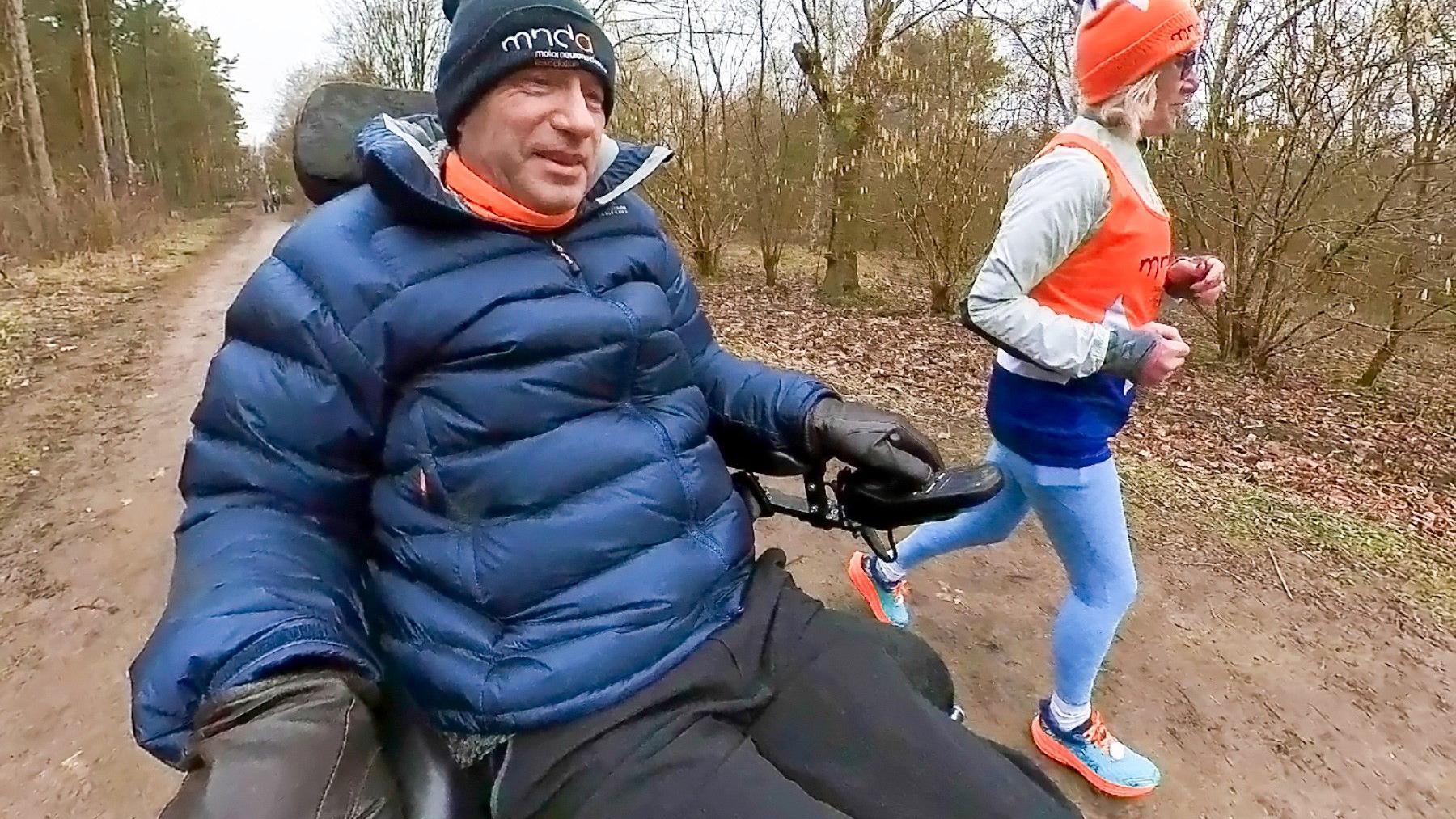 A man in a powerchair, dressed in warm clothes, drives down a country track alongside a jogging woman