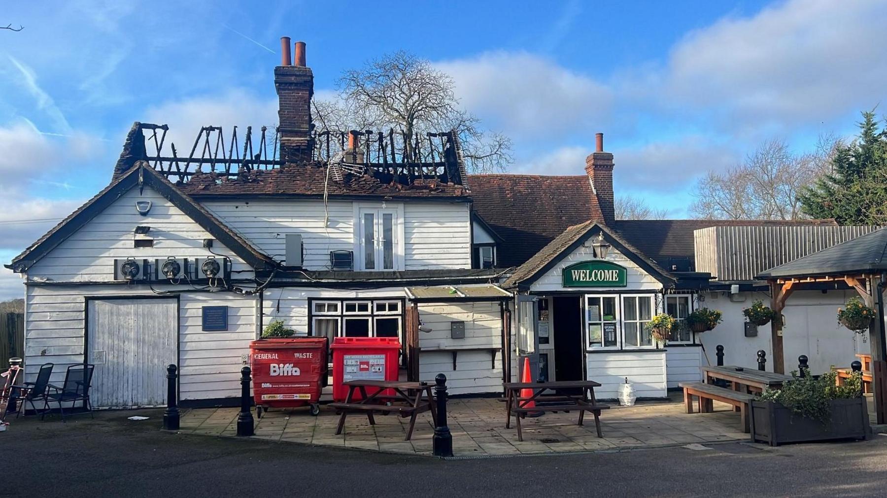 The gutted roof of the pub the following morning. A few smoke-blackened beams are all that remain of a large section of roof. Red bins still stand outside the pub and there are tables and benches outside too. The pub walls is painted white.