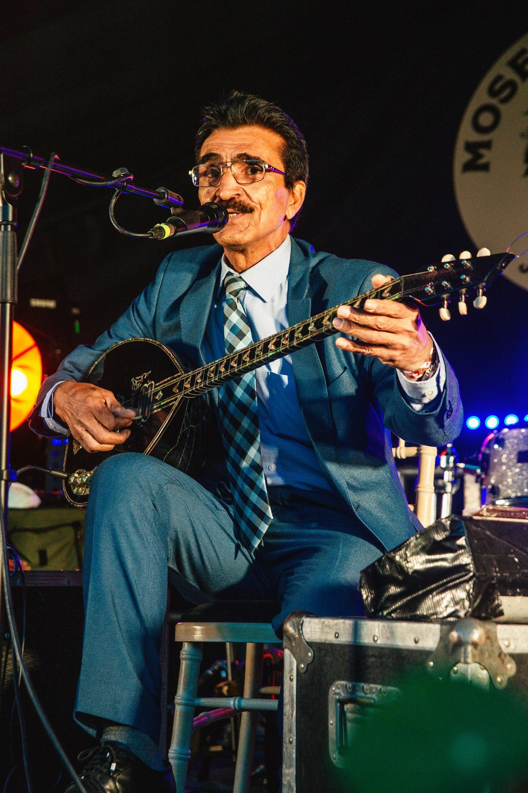 A dark-haired Mohammad Syfkhan wears a suit and checked tie. He is playing a Bouzouki on stage and singing into a microphone.