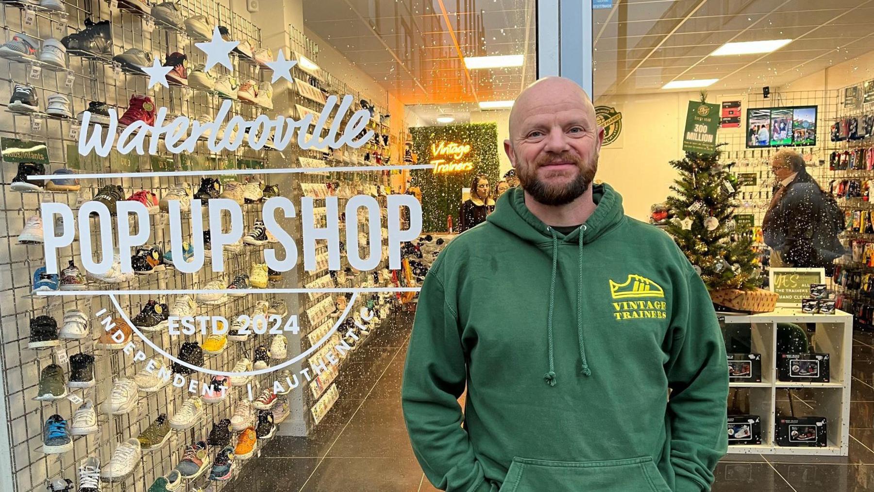 Matt Shaul, in a green Vintage Trainers branded hoodie stood outside the front of the new store which has "Waterlooville pop up shop Est 2024 Independent Authentic" printed on the glass outside. Behind the glass inside the store a man is shopping and there are walls full of shoes.