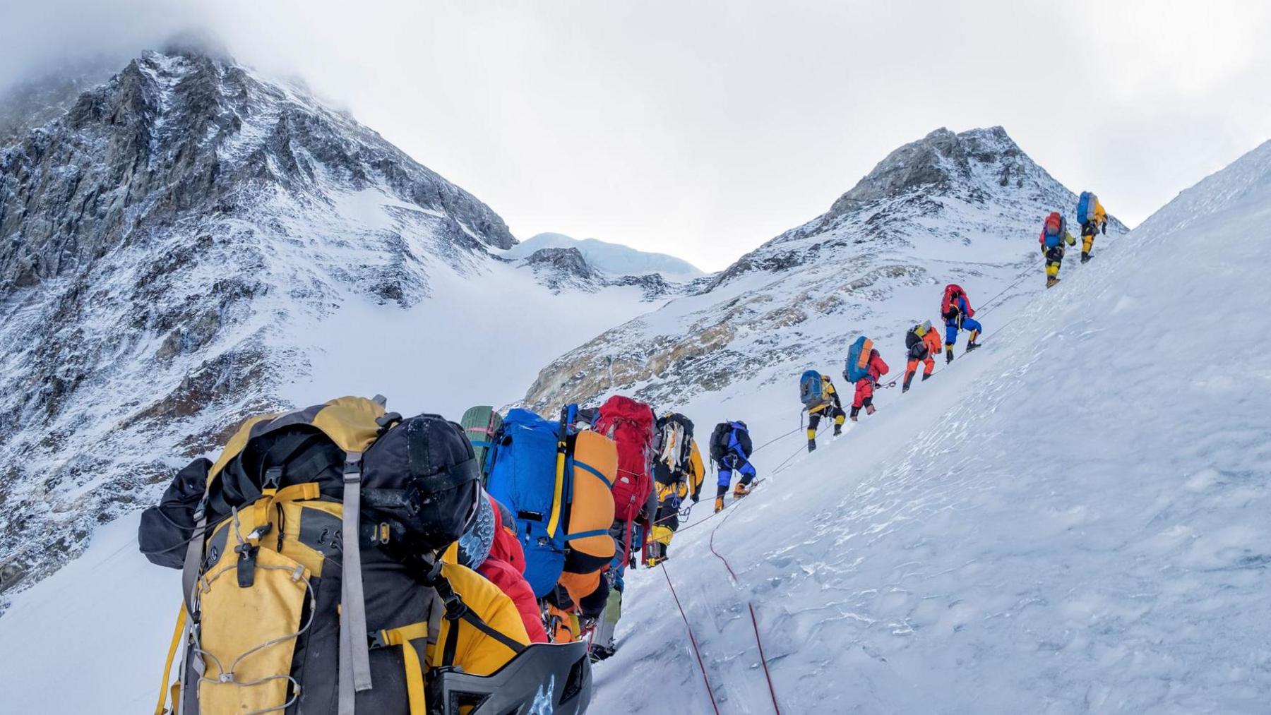 A line of mountain climbers hike across a snow-covered slope with mountain peaks in the background